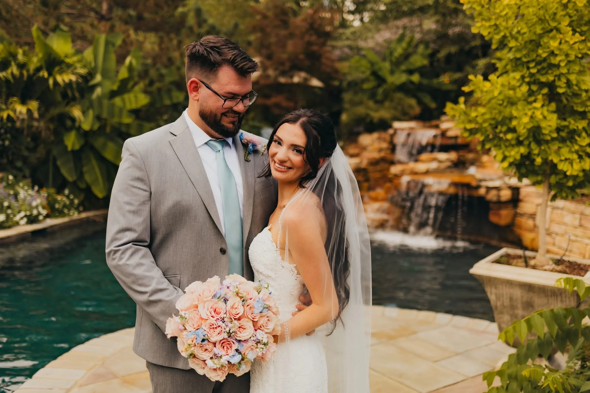 A newlywed couple standing by a pond with a waterfall in the background, holding a bouquet of pastel-colored flowers, smiling at the camera outdoors.