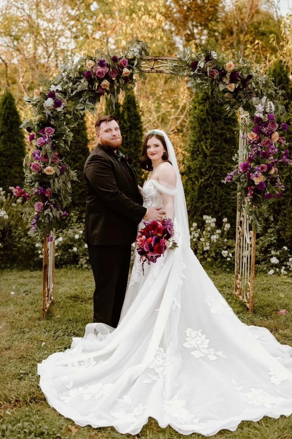 A bride and groom standing close together in front of a floral arch at an outdoor wedding. The bride is holding a bouquet and wearing a white wedding gown with a long train, veil, and lace details. The groom is in a black suit.