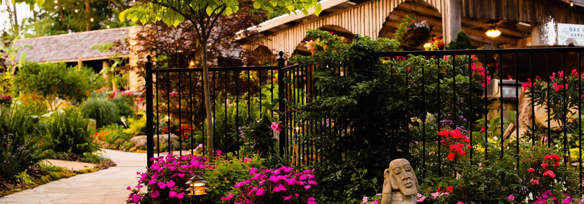 A garden with a stone pathway, vibrant flowers, and lush greenery enclosed by black metal fencing, with a wooden building in the background.