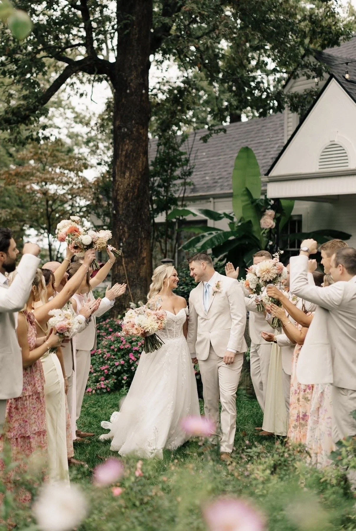 A wedding celebration outdoors with a bride and groom smiling at each other, surrounded by friends and family holding bouquets and raising their hands in celebration during daytime.