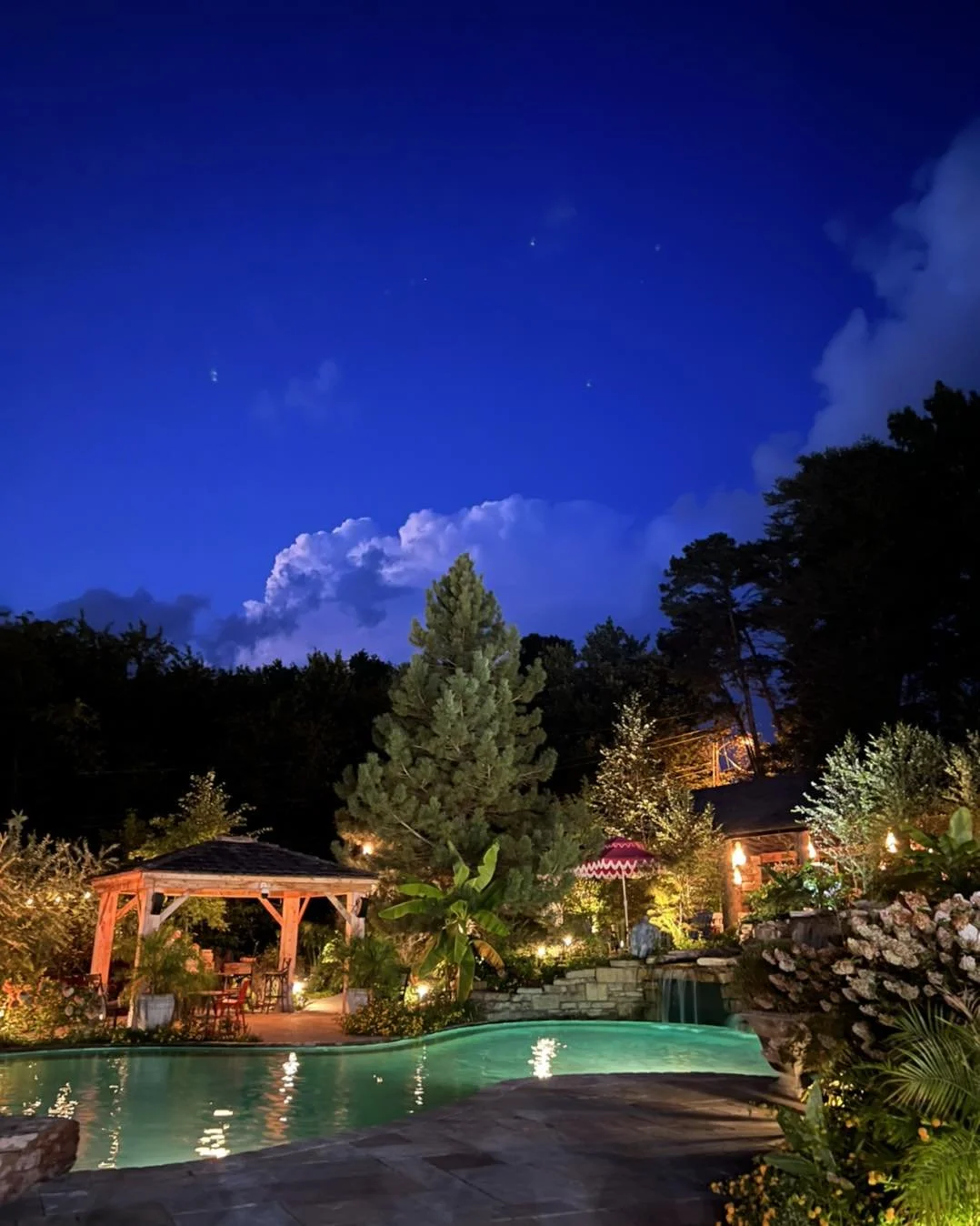 Nighttime scene of a backyard with a lit swimming pool, surrounded by trees and plants, a wooden gazebo, outdoor furniture, and a house with warm lights, under a dark blue sky with clouds and visible stars.