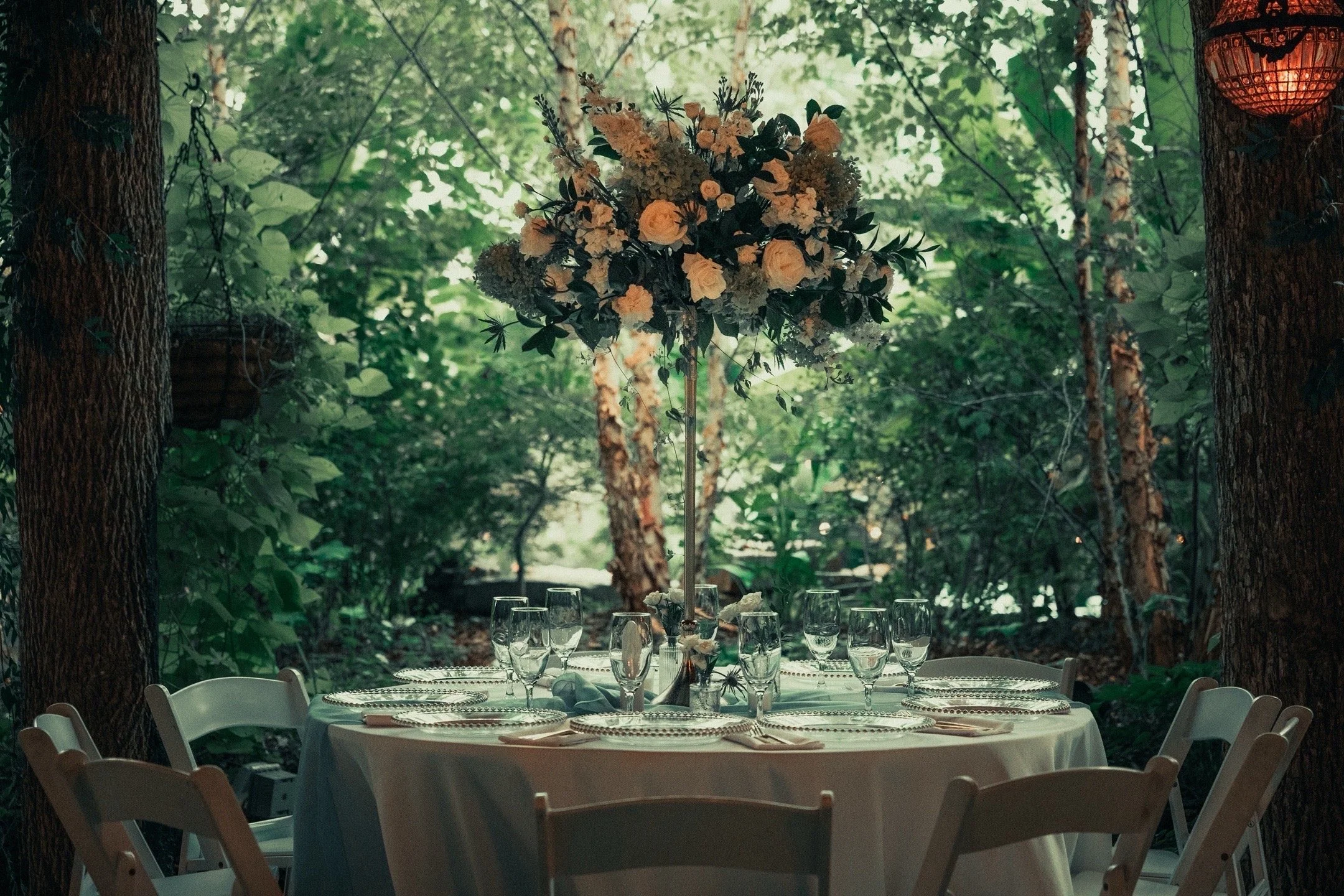 A round banquet table set for a formal event in a forest, with wine glasses, plates, napkins, and a tall floral centerpiece with white roses and greenery, surrounded by white chairs, with trees and green foliage in the background.