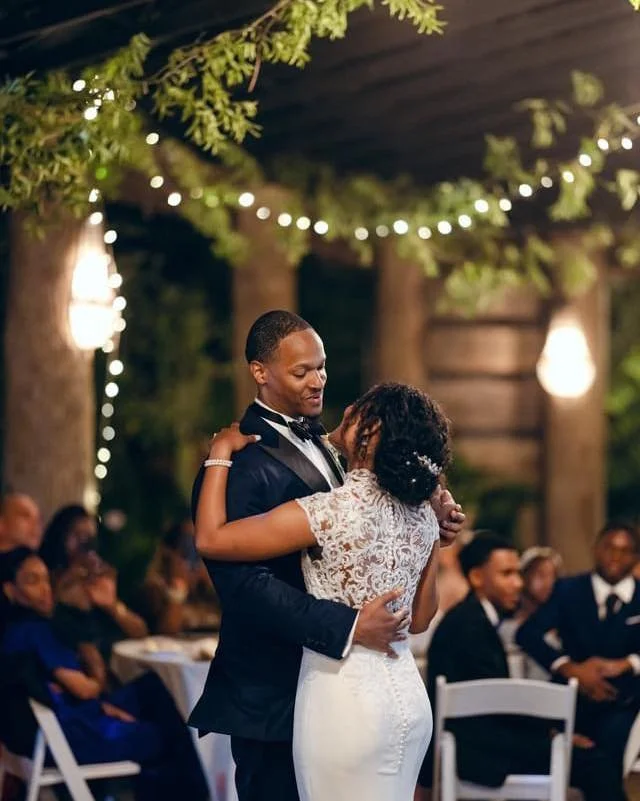 A couple is dancing at their wedding reception outdoors at night, surrounded by guests, with string lights and trees in the background.