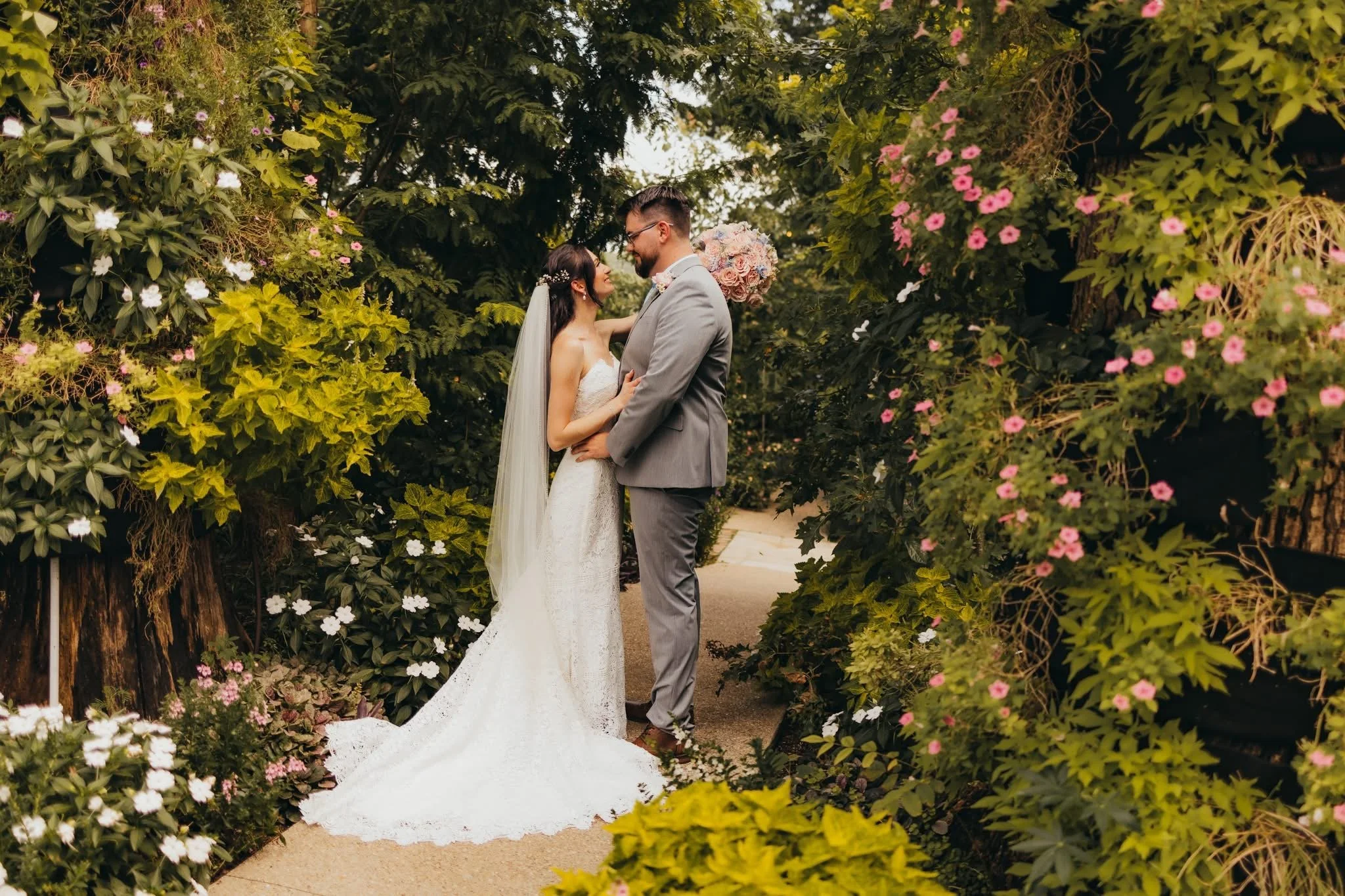 A bride and groom share an intimate moment in a lush, flower-filled garden. The bride wears a white lace wedding dress and veil, while the groom is dressed in a gray suit. They are looking into each other's eyes, surrounded by pink and white flowers 