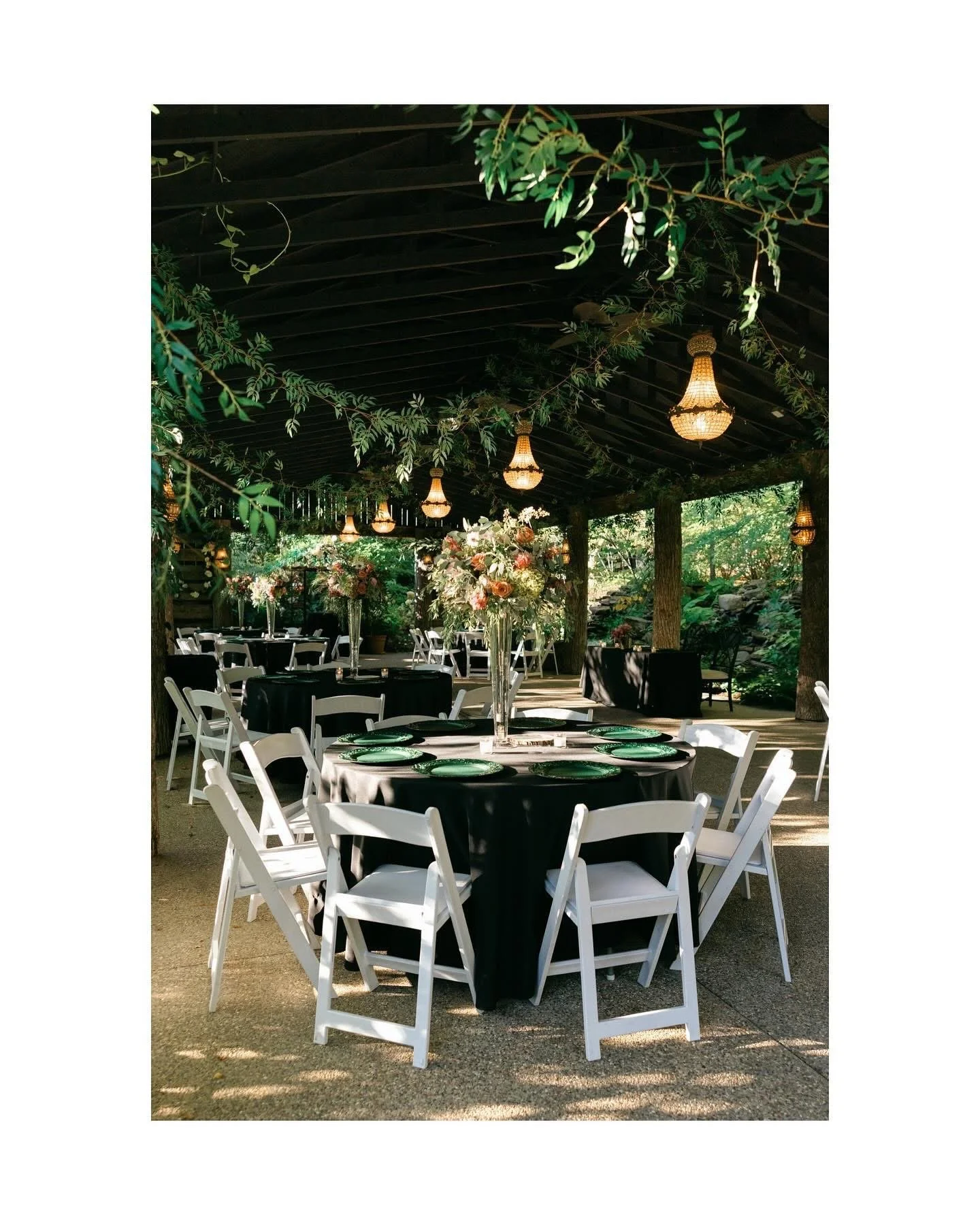 Elegant outdoor event space with round tables draped in black tablecloths, decorated with tall floral centerpieces, surrounded by white chairs, under hanging chandeliers in a lush, green garden setting.