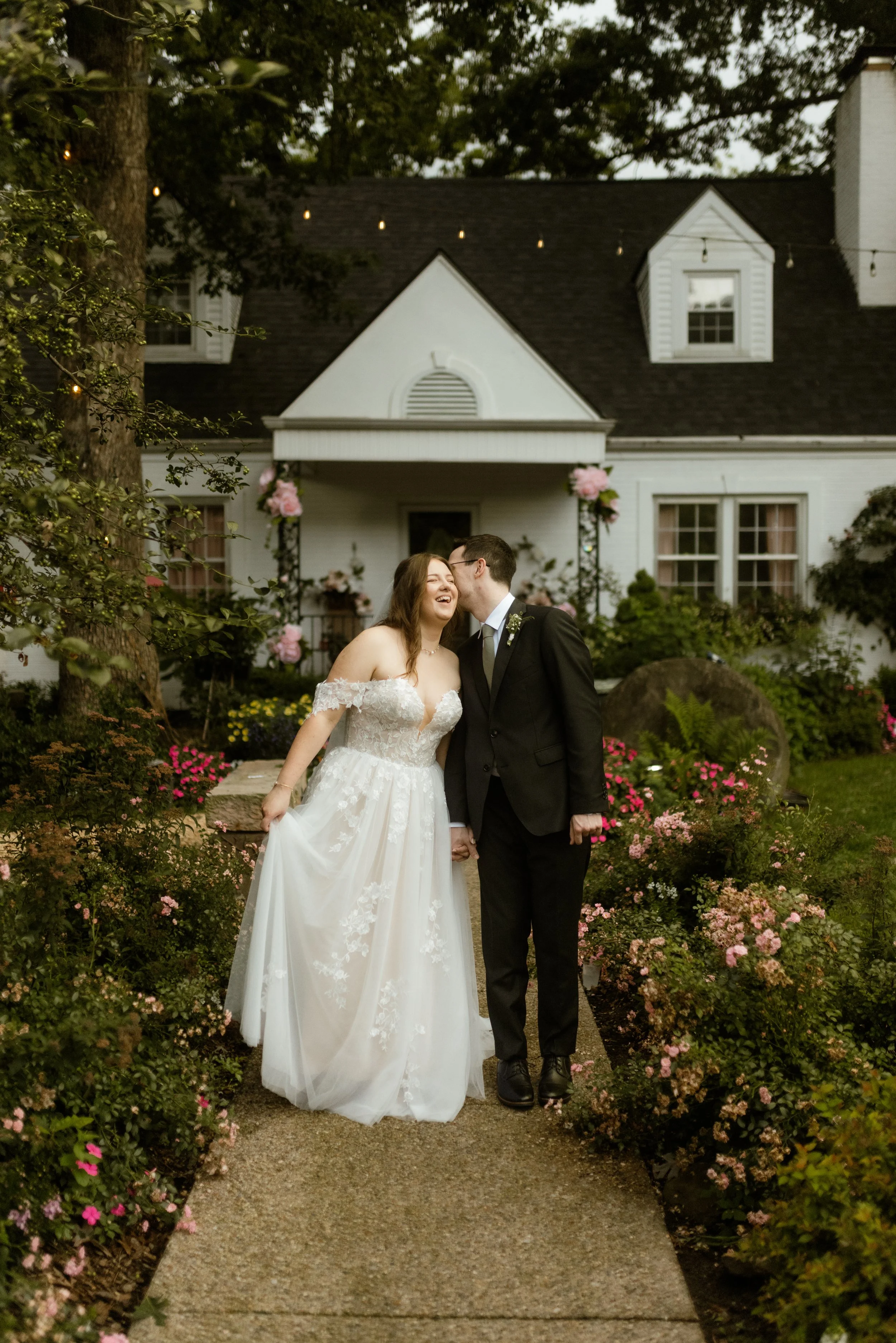 A newlywed couple in wedding attire sharing a kiss in a garden with colorful flowers and greenery.