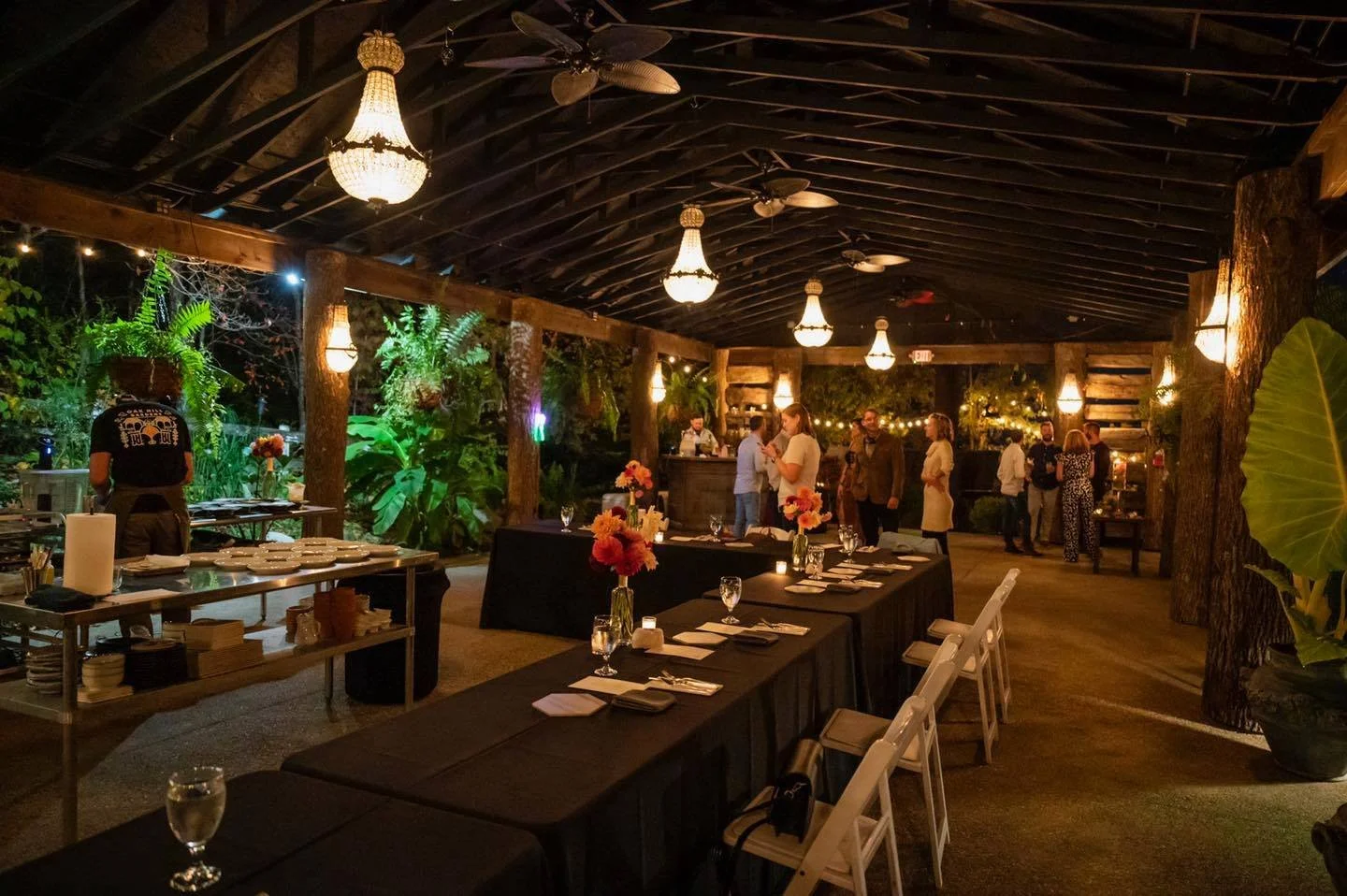 Indoor dining area decorated for an event, with black tablecloths, floral centerpieces, and warm lighting, featuring a bar in the background and a lush outdoor view with plants.