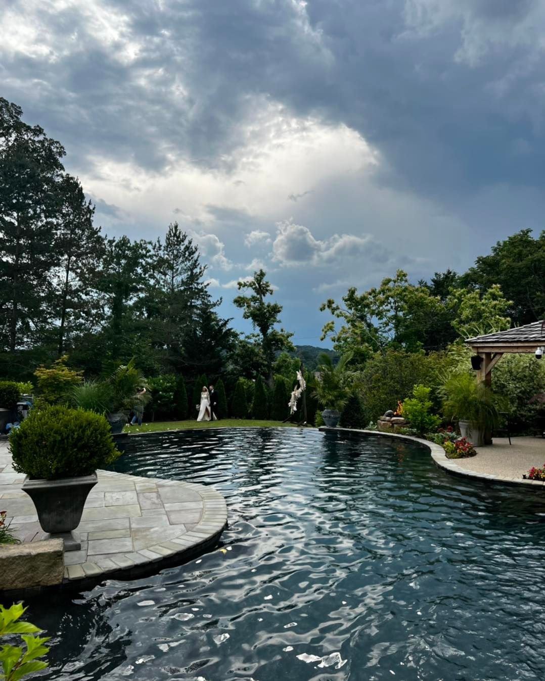 A backyard with a swimming pool surrounded by trees and plants, a cloudy sky overhead, and two people dressed in white attending a wedding.
