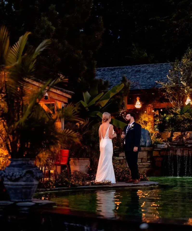 A bride and groom standing by a pond during a wedding reception at night, surrounded by lush greenery and warm lighting.