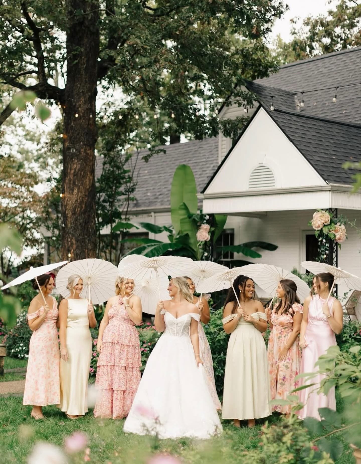 A bride in a white wedding gown and six bridesmaids in pastel dresses holding white umbrellas on a garden lawn for a wedding ceremony