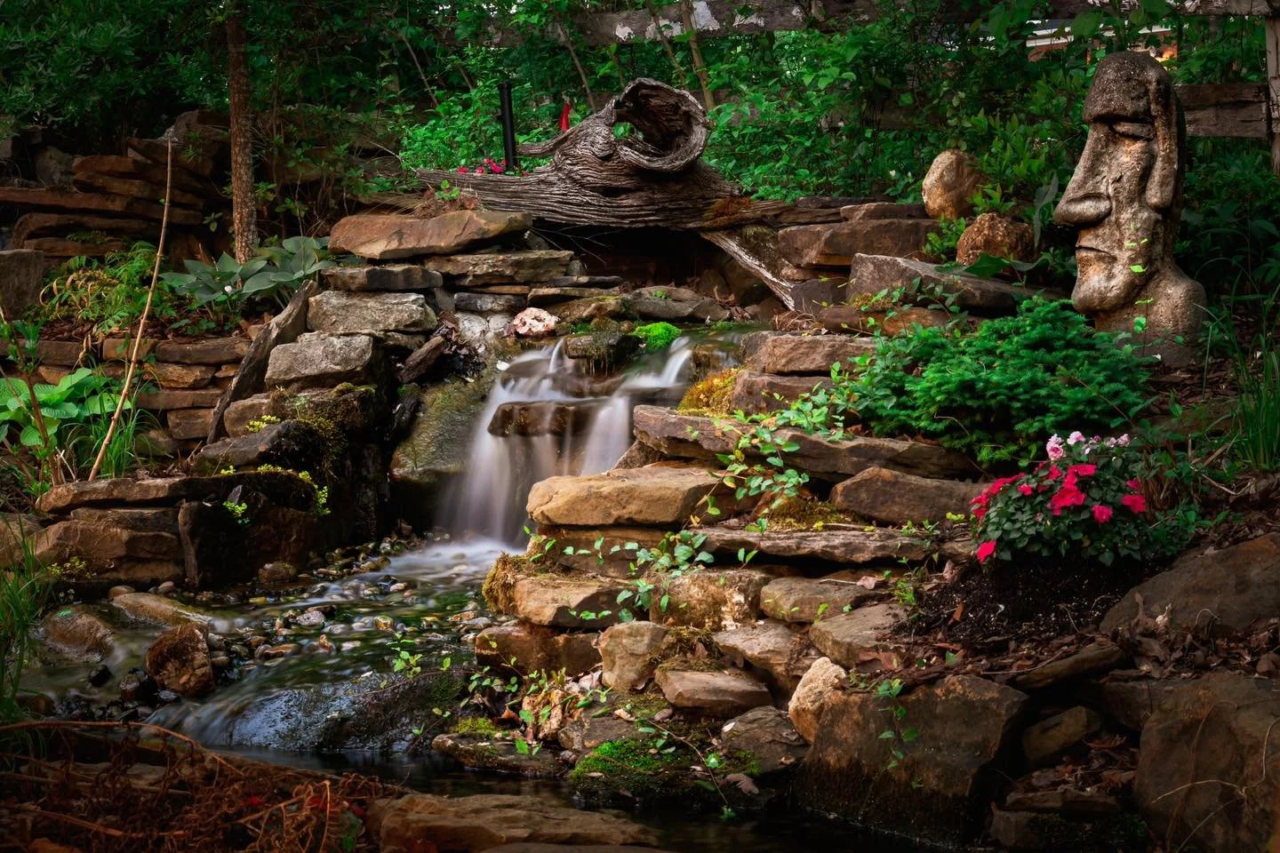 A garden scene with a small waterfall flowing over rocks, surrounded by green plants, flowers, a wooden driftwood piece, and a stone sculpture of a human face.