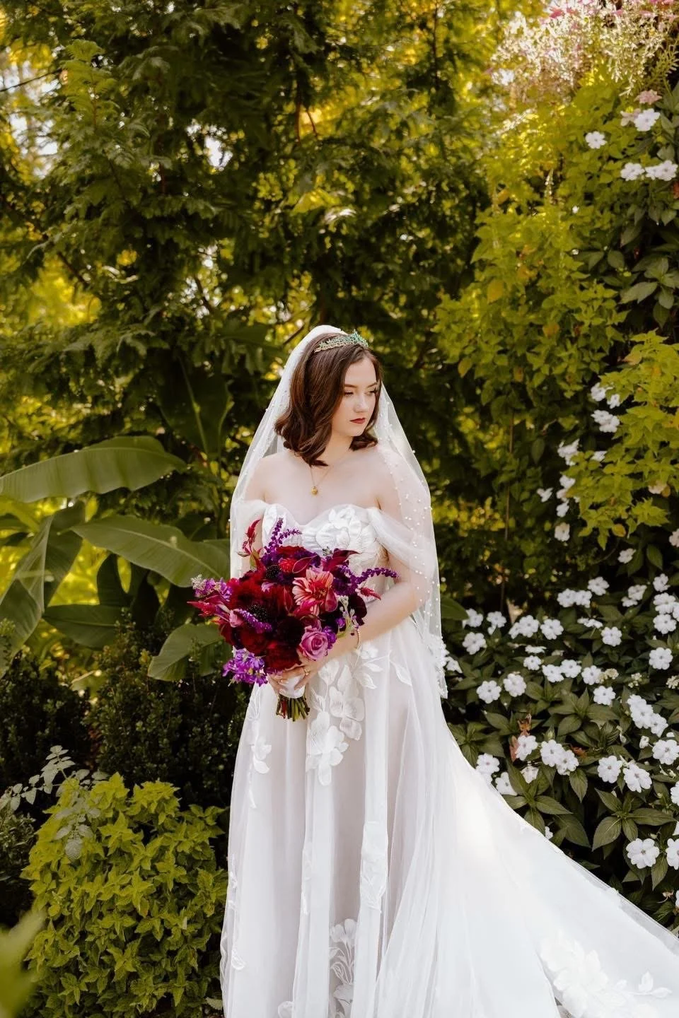 A bride in a white wedding dress holding a bouquet of purple, red, and pink flowers in a lush garden.
