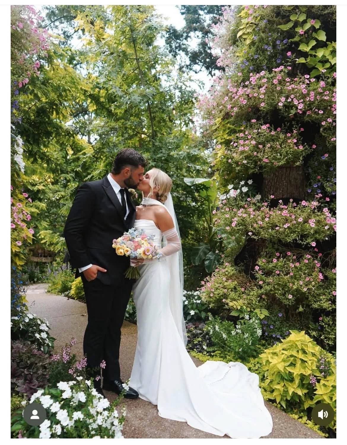 A bride and groom share a kiss in a lush, flowered garden. The bride is in a white wedding gown holding a bouquet, and the groom is in a black suit with a white shirt. Tucked away in the background are various plants and flowers, creating a romantic 