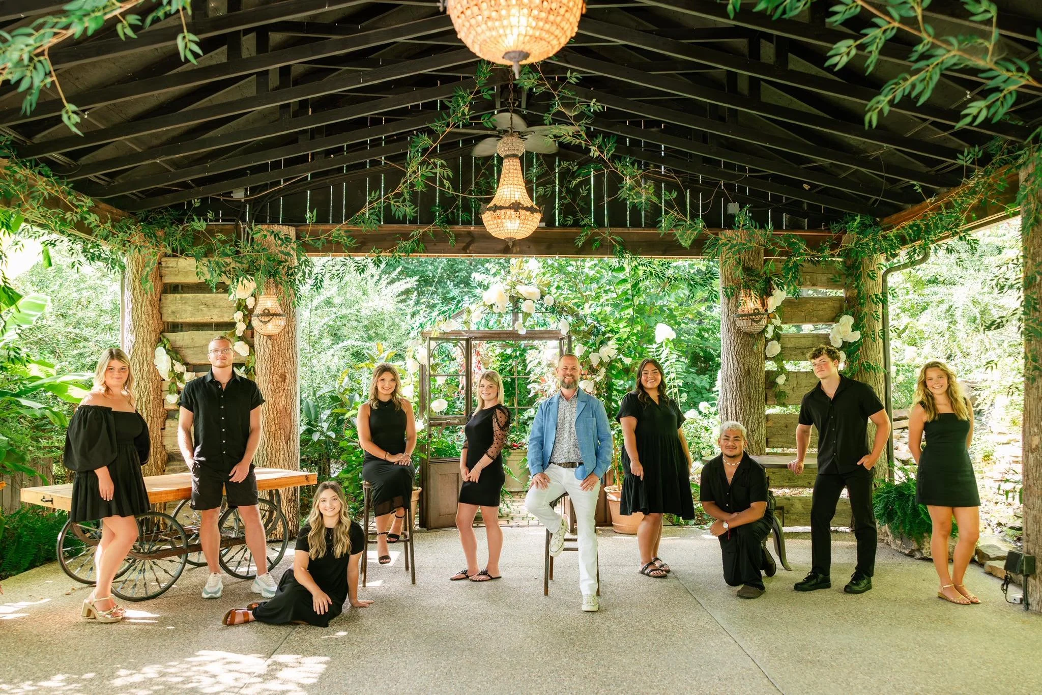 A group of eleven people, seven women and four men, posing inside a lush, green botanical garden with wooden structures and hanging lights.