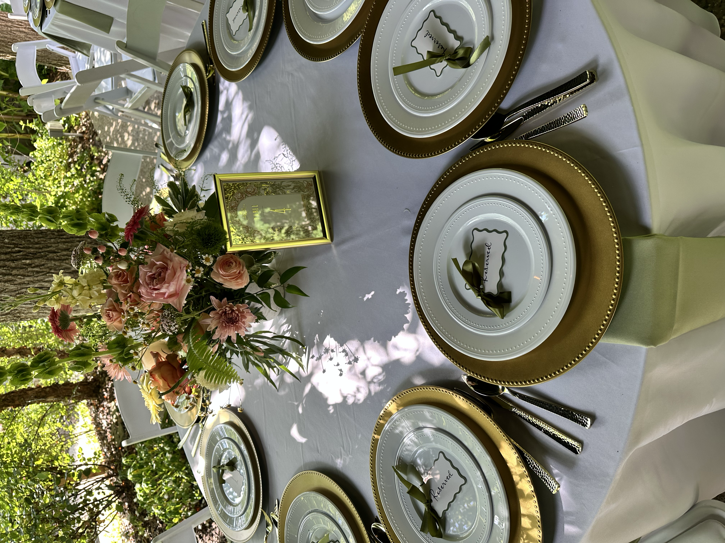 Elegant outdoor table setting with white tablecloth, gold-trimmed white plates, green napkins, and silverware. A floral centerpiece with pink roses, yellow flowers, and green foliage. Small framed signs on the table and a natural background with tree