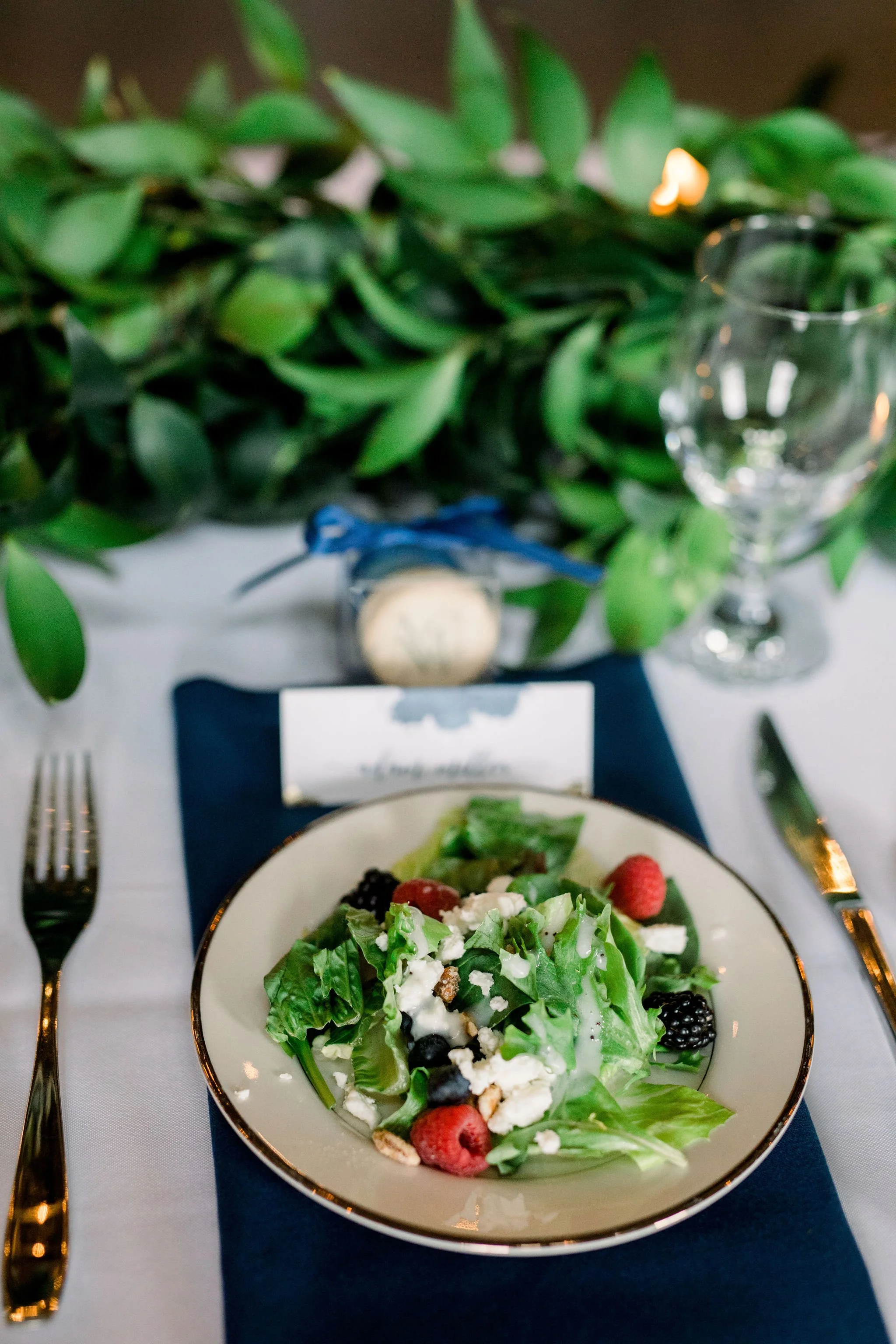 A plate of fresh salad with mixed greens, berries, blue cheese, and nuts on a white tablecloth, set with gold utensils, a blue napkin, and a glass of water, with green foliage and a decorative candle in the background.
