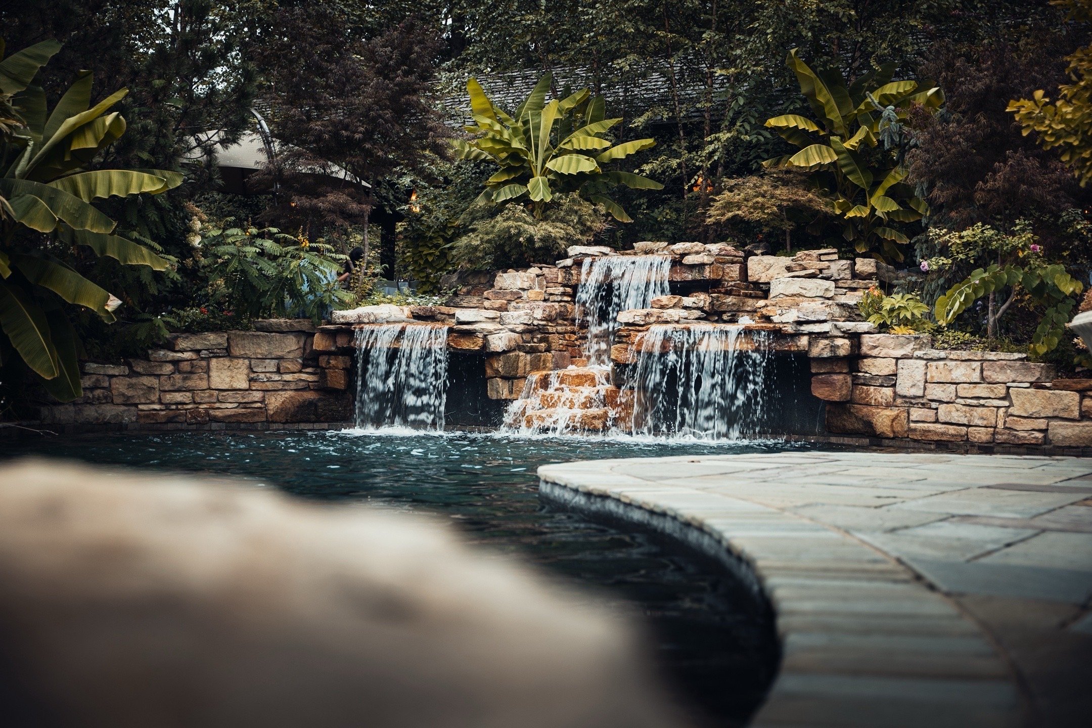 A backyard with a stone waterfall feature flowing into a swimming pool, surrounded by lush green plants and trees.