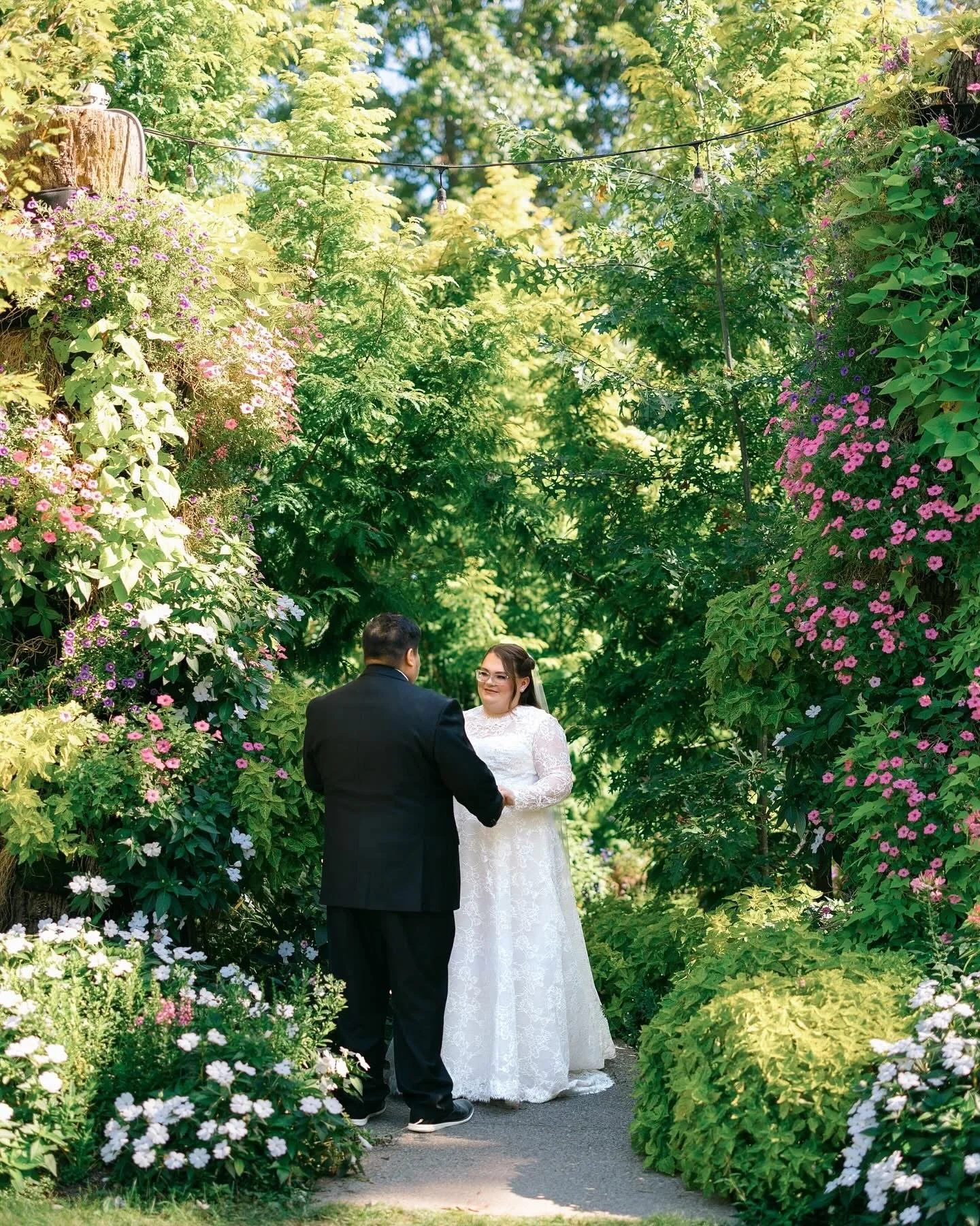 A couple dressed in wedding attire holding hands in a garden with lush green trees and colorful flowers.