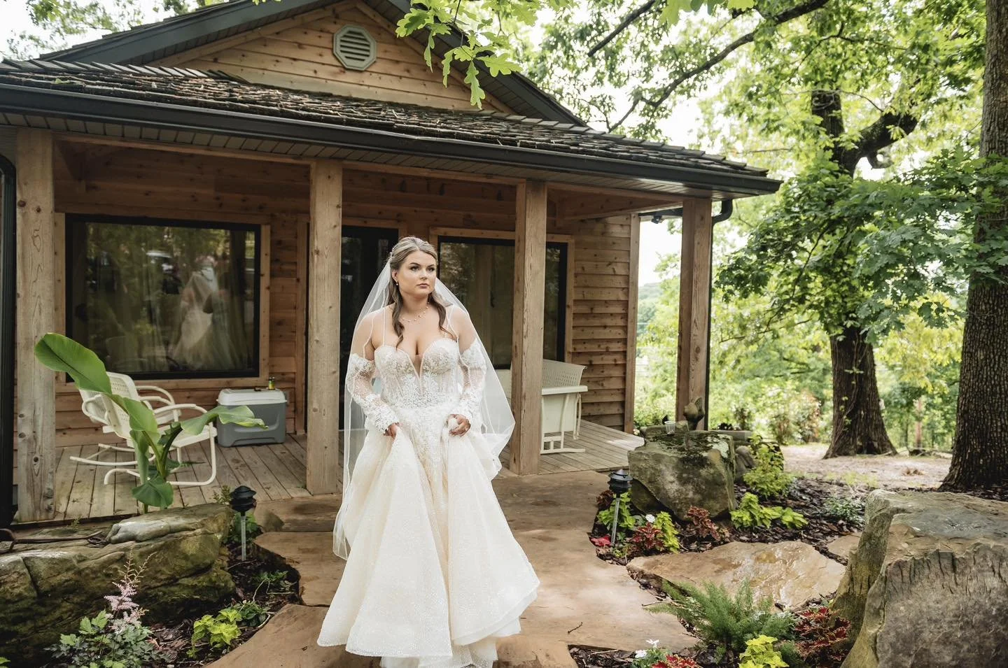 A bride in a wedding dress standing on a wooden porch of a house surrounded by greenery and trees.