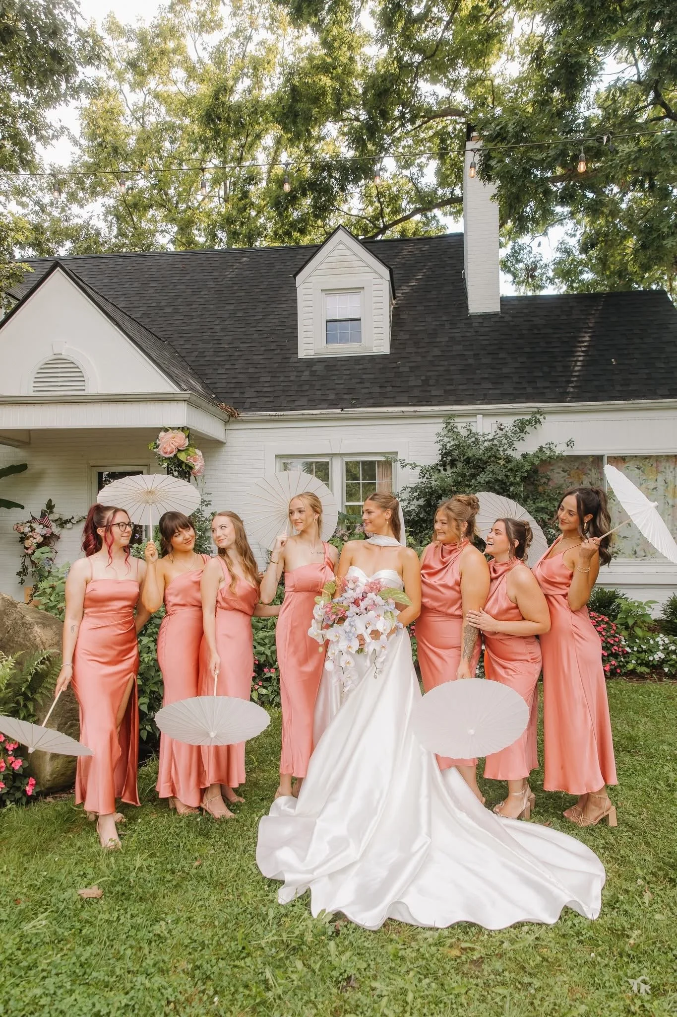 A bride in a white wedding dress holding a bouquet of flowers standing in front of bridesmaids in matching satin pink dresses, posing outdoors in a garden with greenery and a white house with black roof in the background. The bridesmaids are holding 