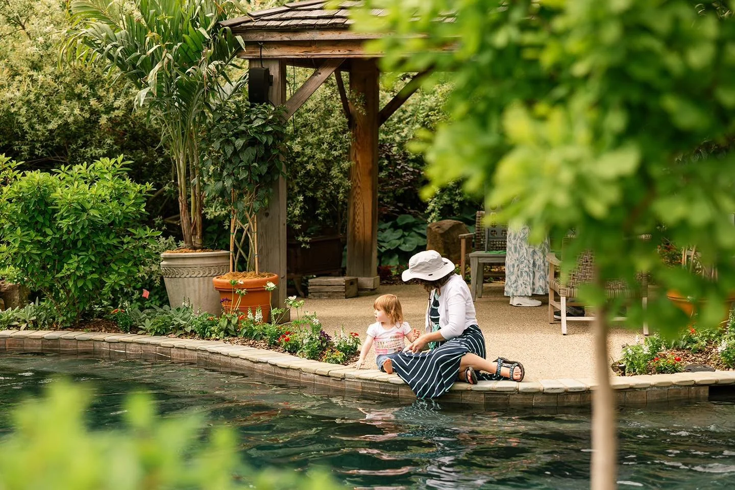 A woman with a wide-brimmed white hat and a child sitting by a pond, touching the water, surrounded by lush green plants and garden furniture.