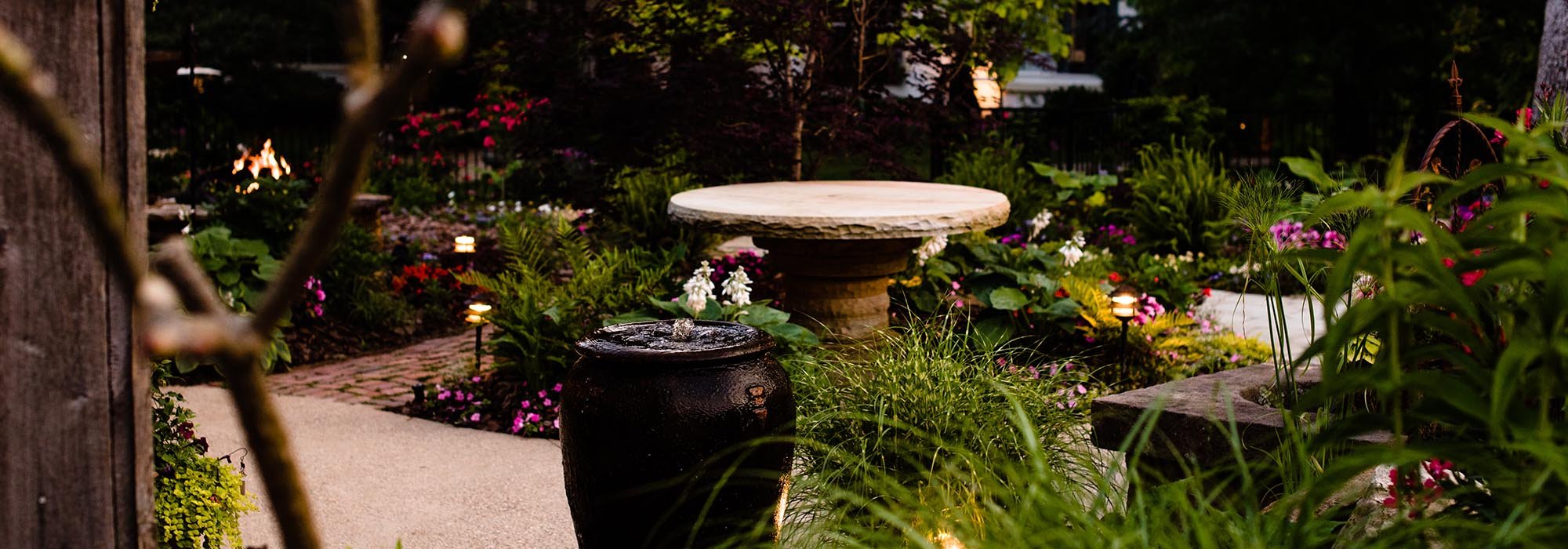 A well-lit garden with a stone birdbath surrounded by colorful flowers and greenery, evening setting.