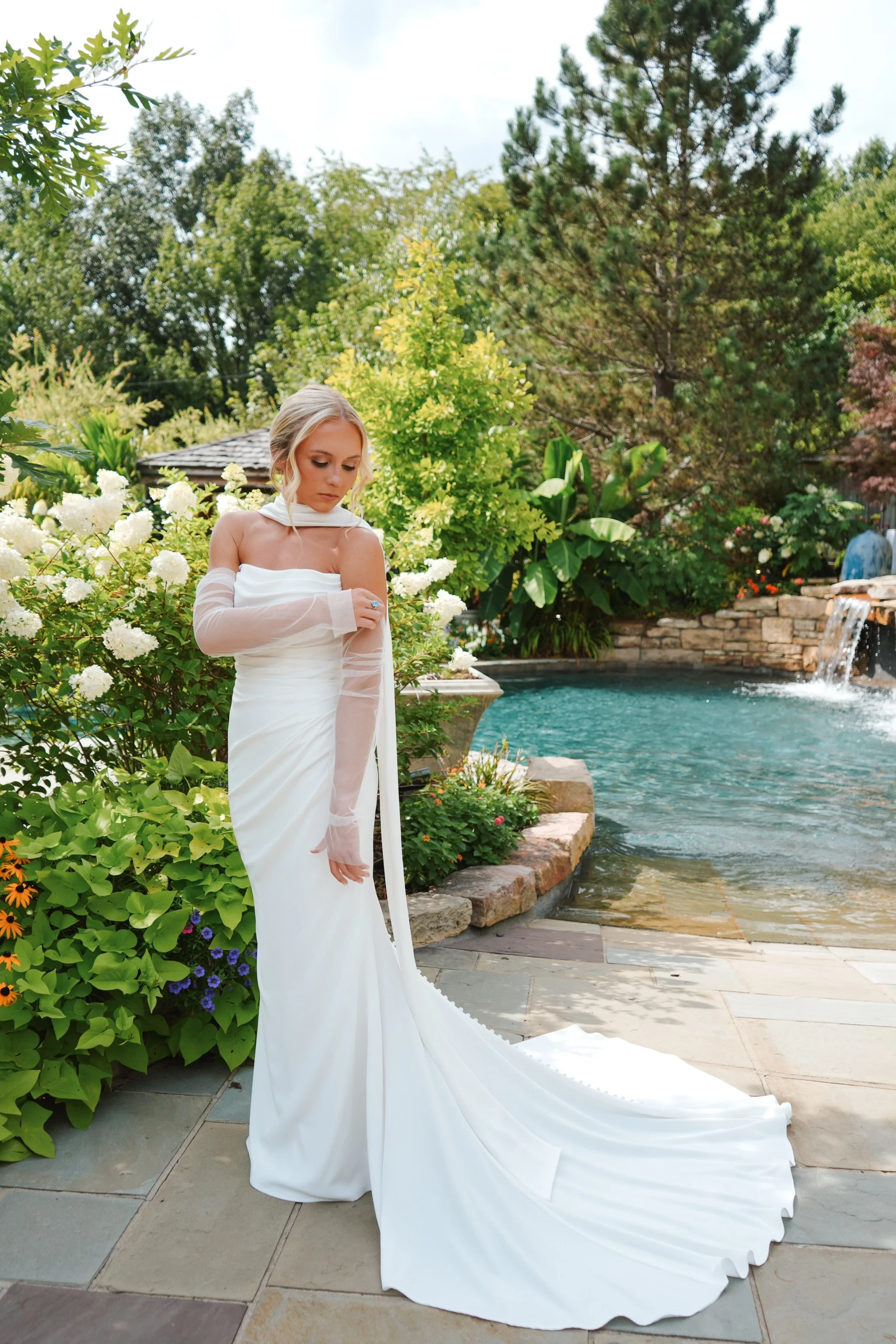 A woman in a white wedding dress standing on a stone patio near a pool and garden, with trees and flowers in the background.