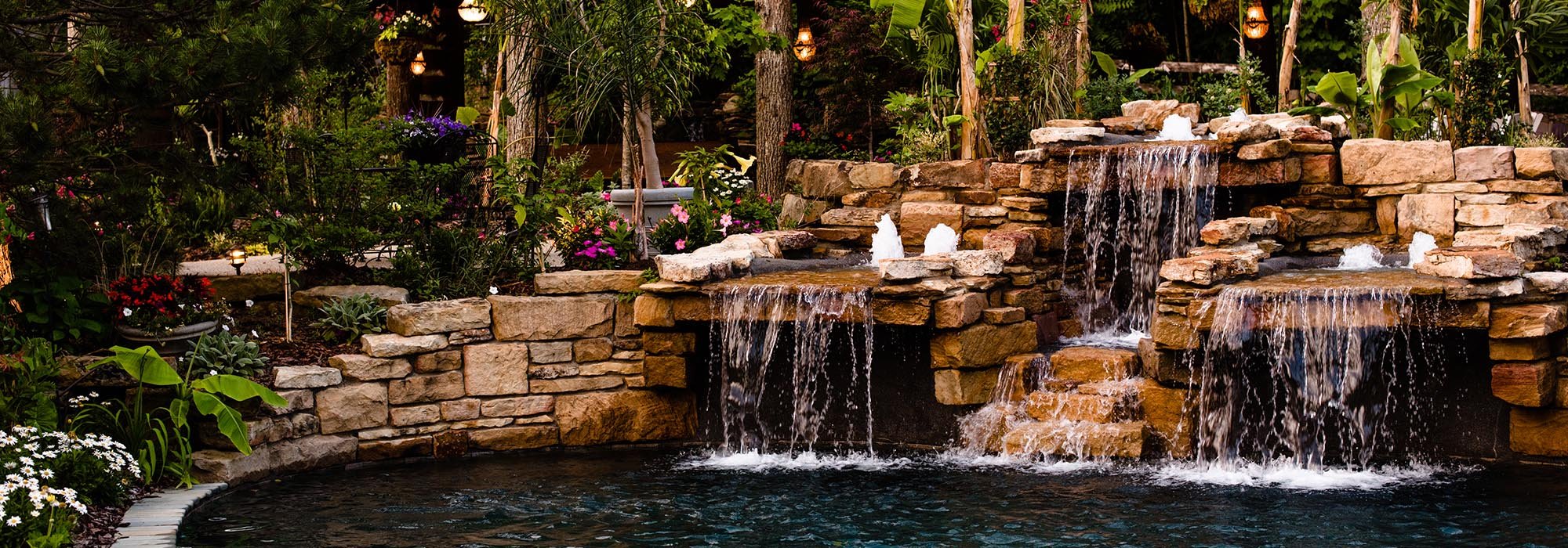 A backyard garden with a stone waterfall feature flowing into a pond, surrounded by lush plants, flowers, and trees.