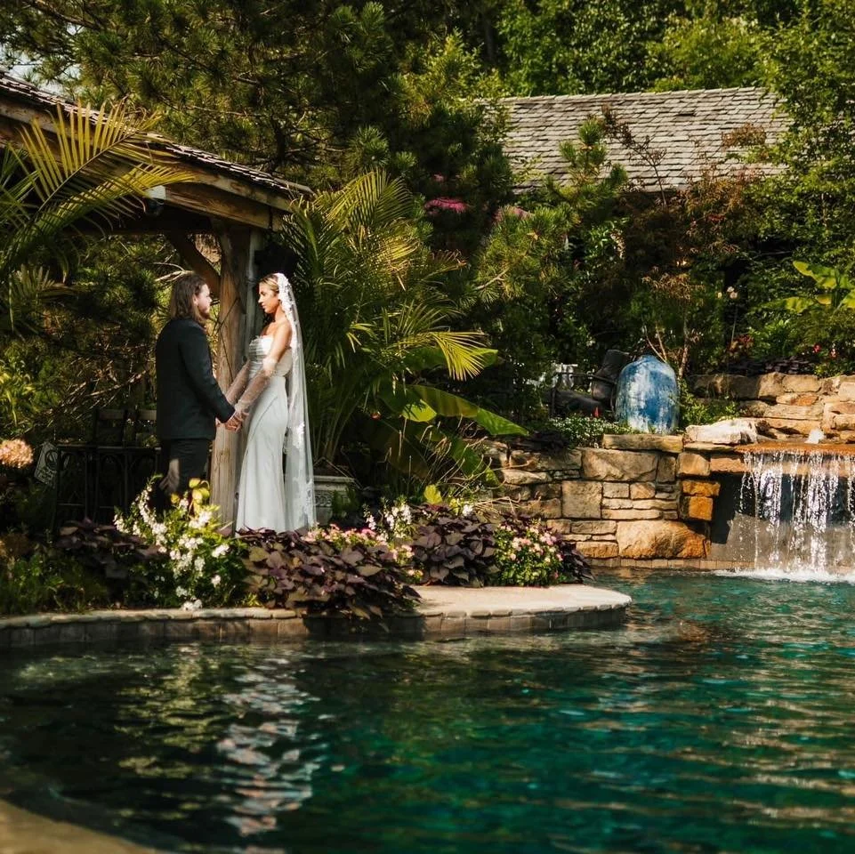A bride and groom standing hand in hand near a pool, surrounded by lush greenery and rocks, during their wedding ceremony.