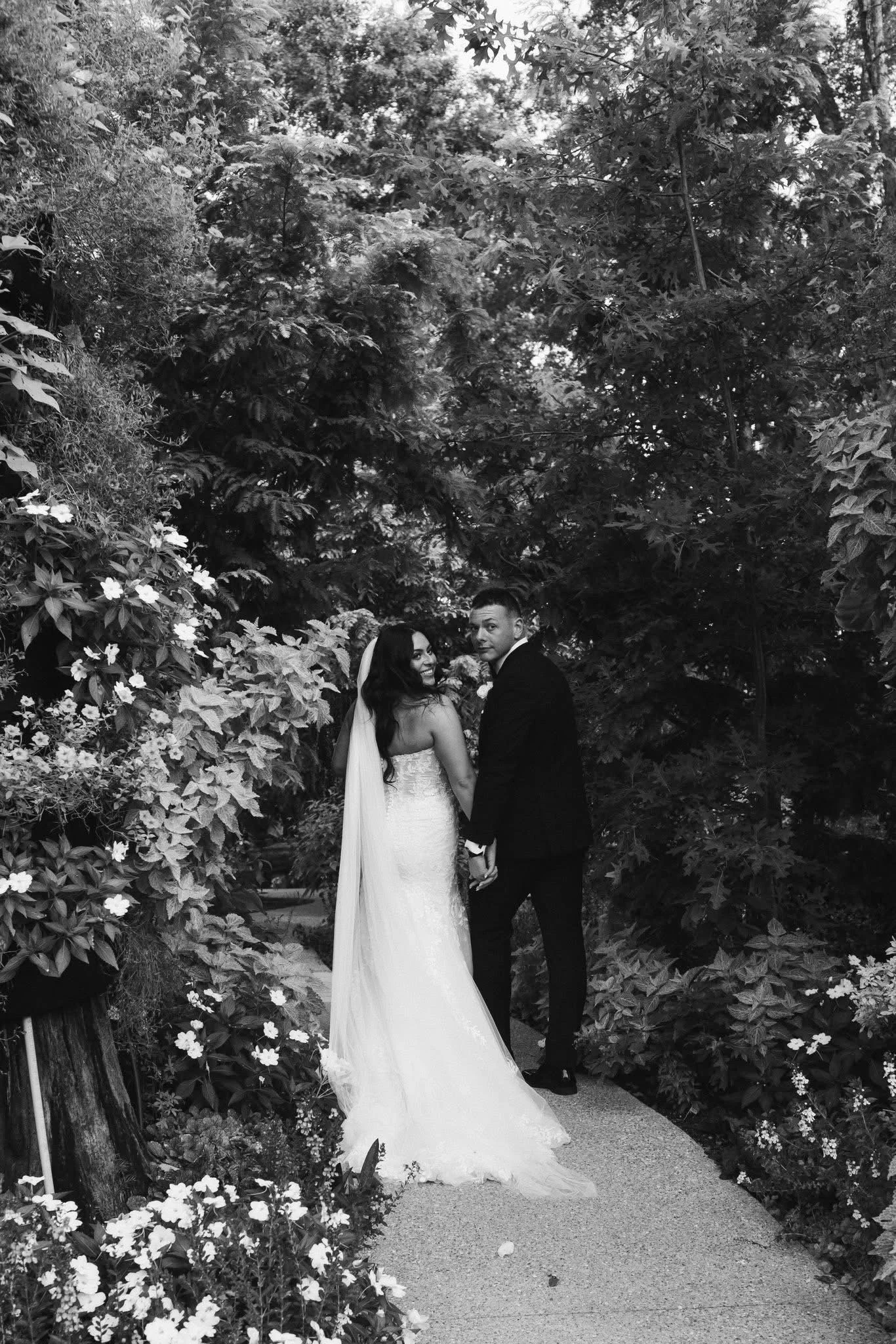 Couple in wedding attire holding hands on a garden pathway surrounded by lush foliage and flowers, smiling at each other.