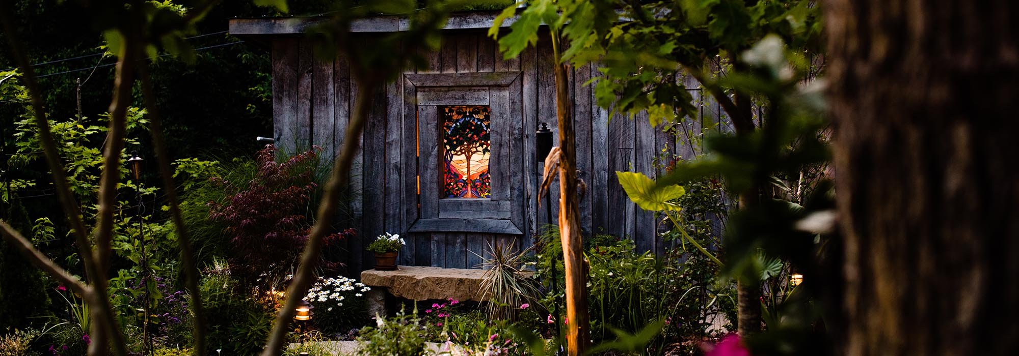 A garden scene with a rustic wooden shed featuring a stained glass window depicting a sunset and trees, surrounded by various plants and flowers, with soft lighting.