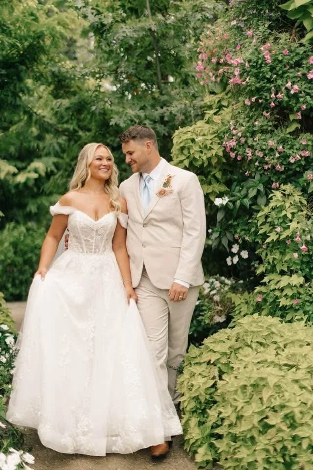 A bride and groom walking together outdoors in a garden, smiling at each other. The bride wears a white wedding gown with off-the-shoulder sleeves, and the groom wears a light-colored suit with a tie and boutonniere. Green foliage and pink flowers su