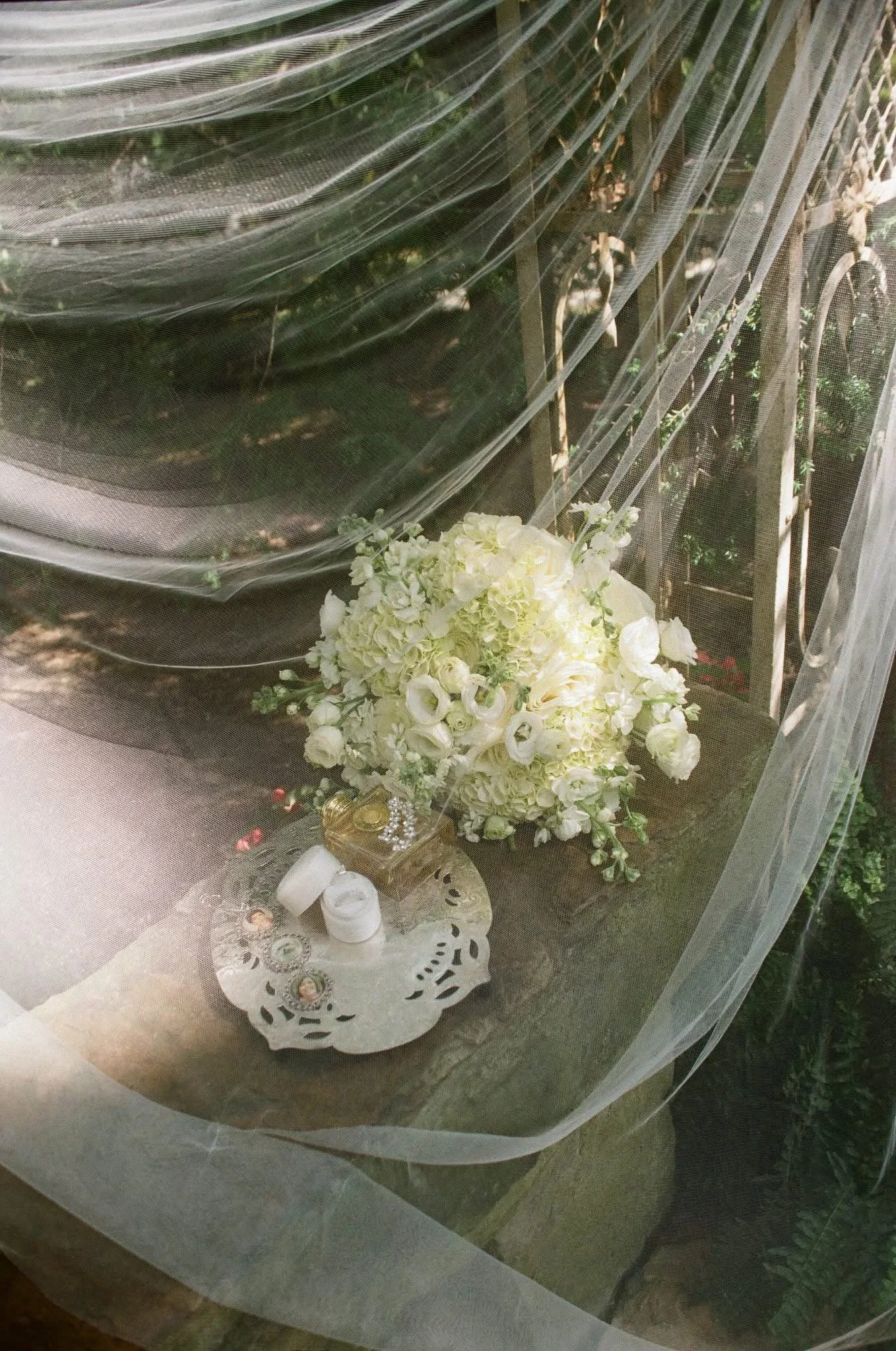 A window with sheer curtains showing a floral arrangement of white flowers on a wooden table outside.