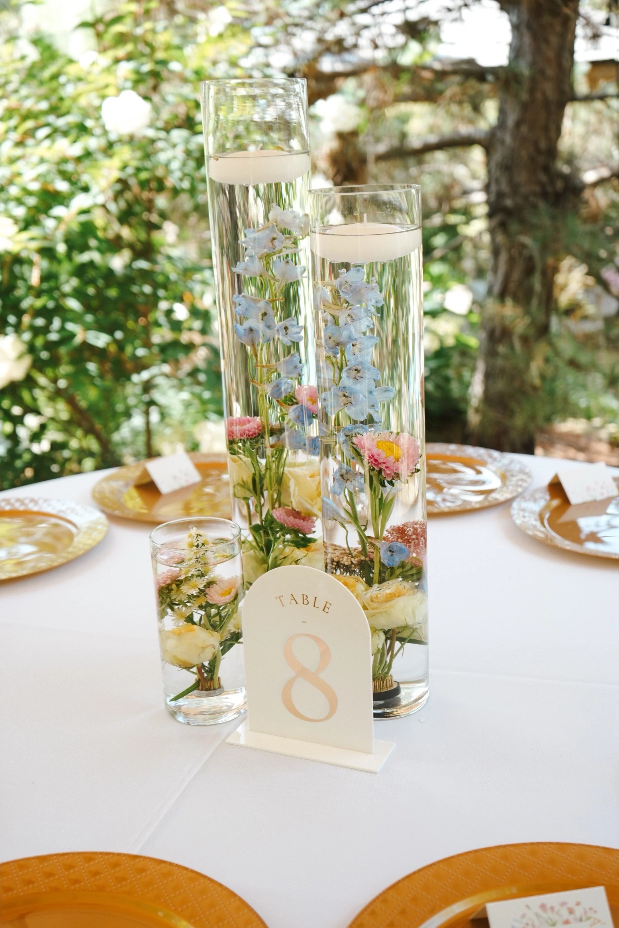 Elegant table centerpiece with tall glass vases filled with floating candles and decorated with blue and pink flowers at a wedding reception table numbered 8.