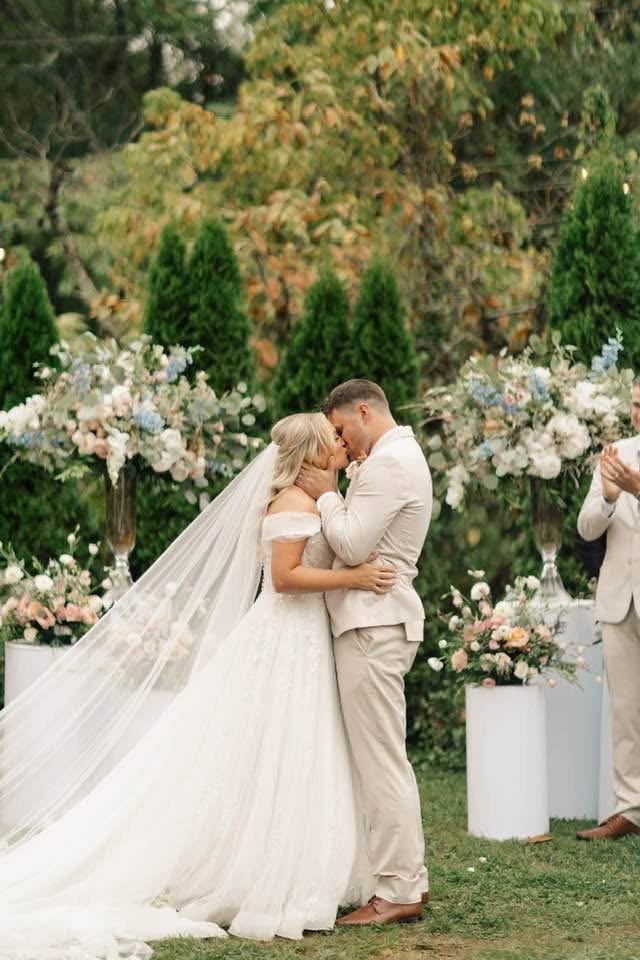 A bride and groom softly touching foreheads during their wedding ceremony outdoors, surrounded by floral decorations and greenery.