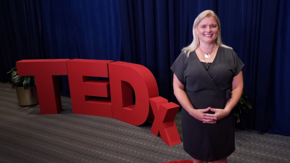 A smiling woman standing next to large red TEDx letters on a stage with a blue curtain background.