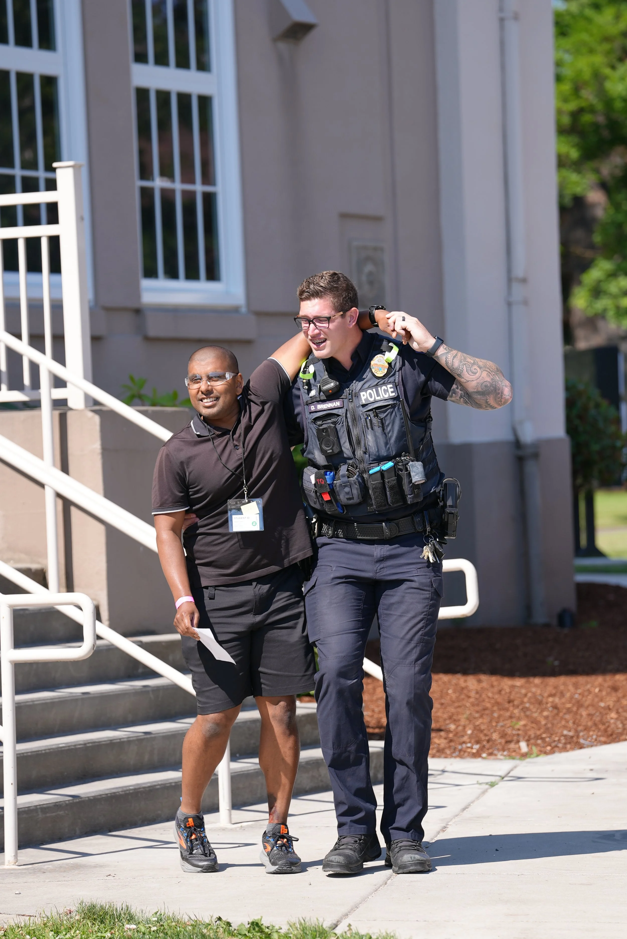 A police officer helping a person with a disability walk up stairs outside a building on a sunny day.