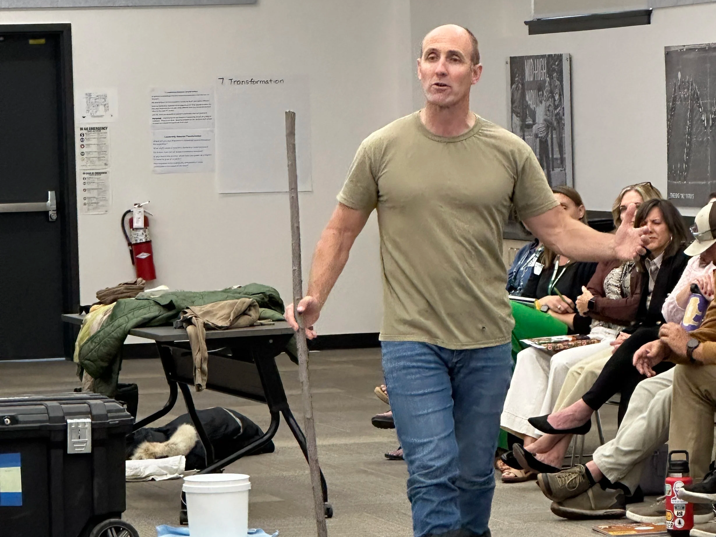 A man in a beige T-shirt and jeans demonstrating walking with oversized boots and a stick in a classroom or lecture hall, with seated audience members watching.