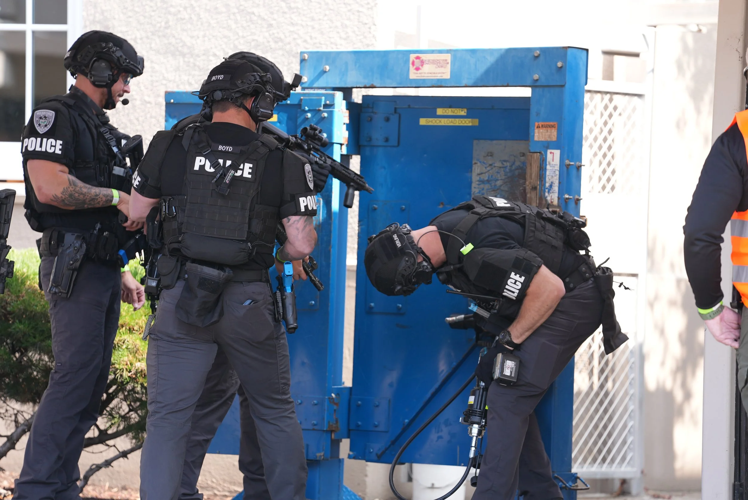 A group of police officers in tactical gear investigating a blue large electrical box outside a building.