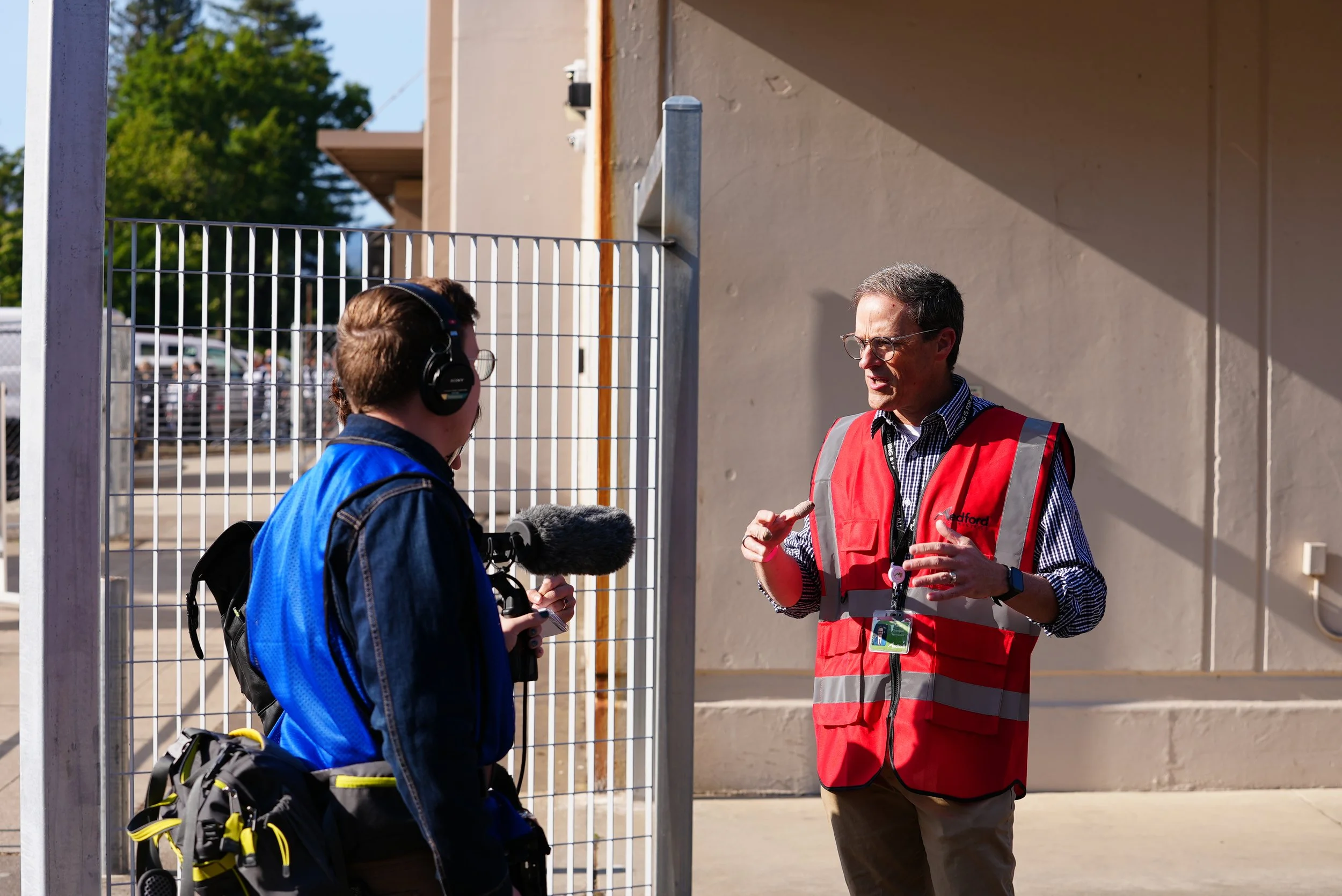 A man in a red safety vest talking to a young man with headphones, a backpack, and a microphone, outside near a beige building and a metal fence.