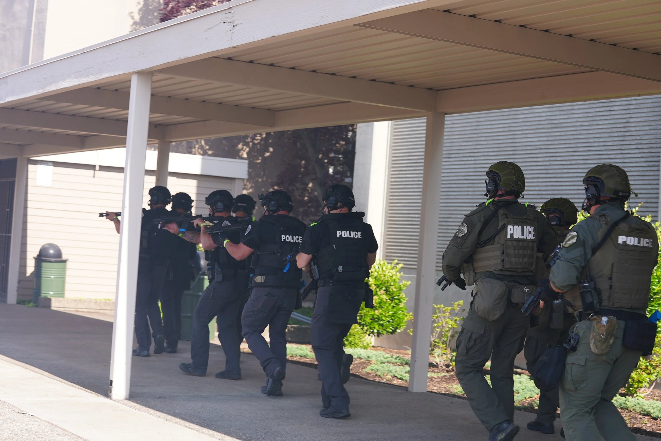 Group of police officers wearing tactical gear and helmets, standing under a sheltered area, aiming weapons during a training or operation.
