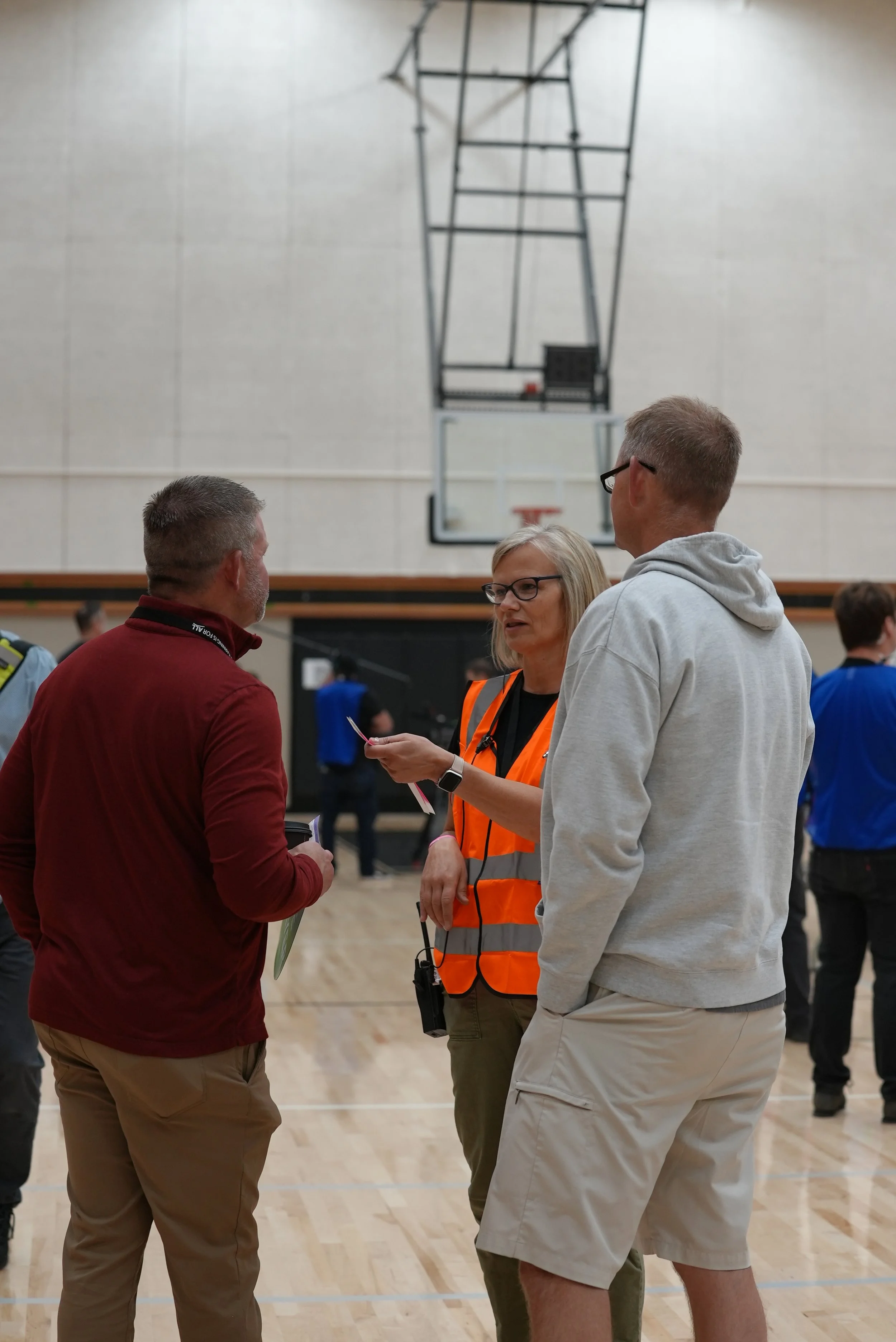 Three people are talking in a gymnasium, with a basketball hoop, on a wooden floor. Two men and a woman, the woman wearing a high-visibility orange vest, are engaged in conversation.