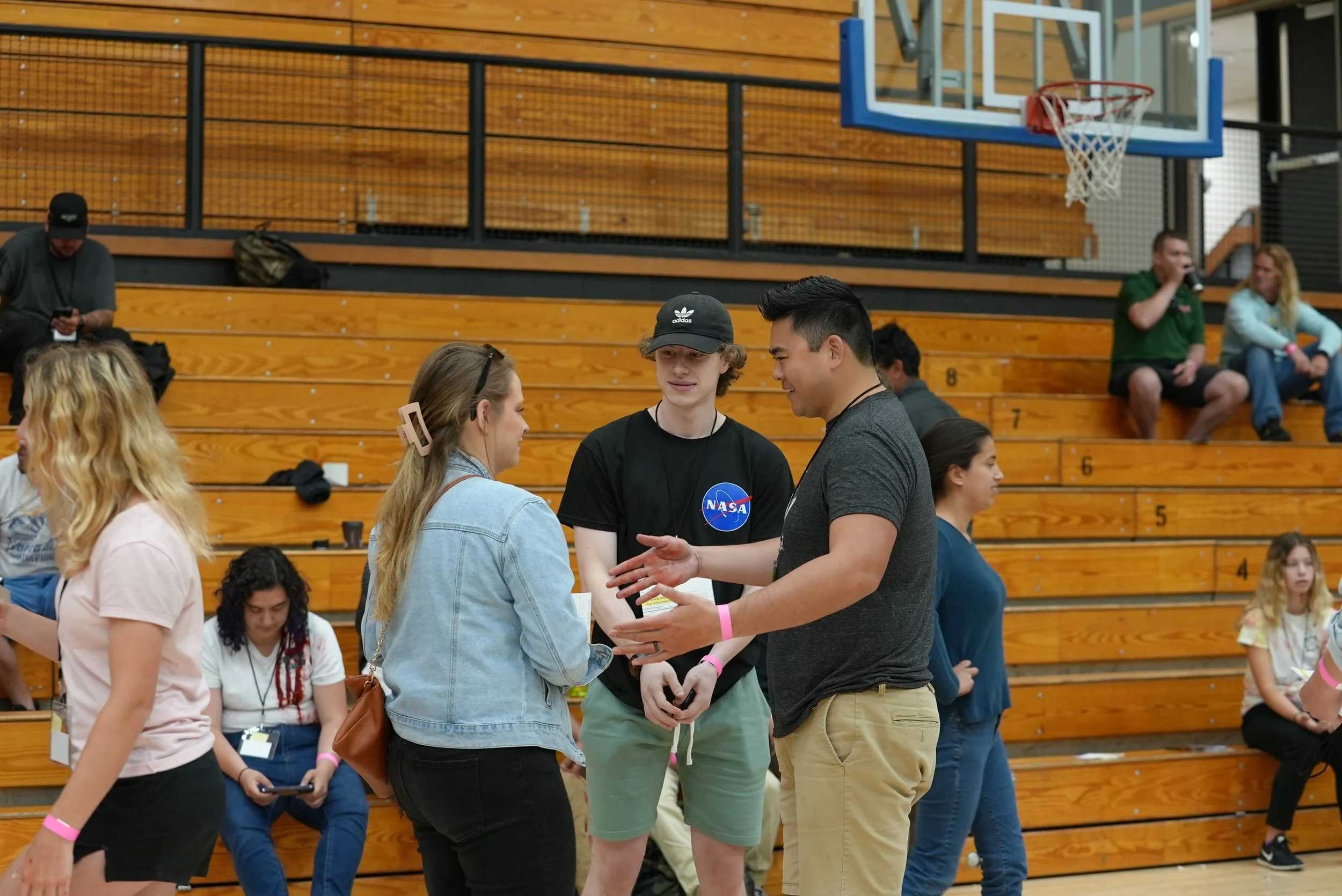 A group of people standing and sitting in a gymnasium with wooden bleachers. Three individuals are engaged in conversation in the foreground, while others sit or stand in the background.