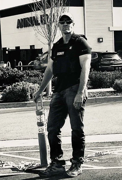 A security guard standing in a parking lot next to a fire extinguisher, wearing sunglasses, a black security shirt, and dark pants, with a building labeled Animal Care in the background.