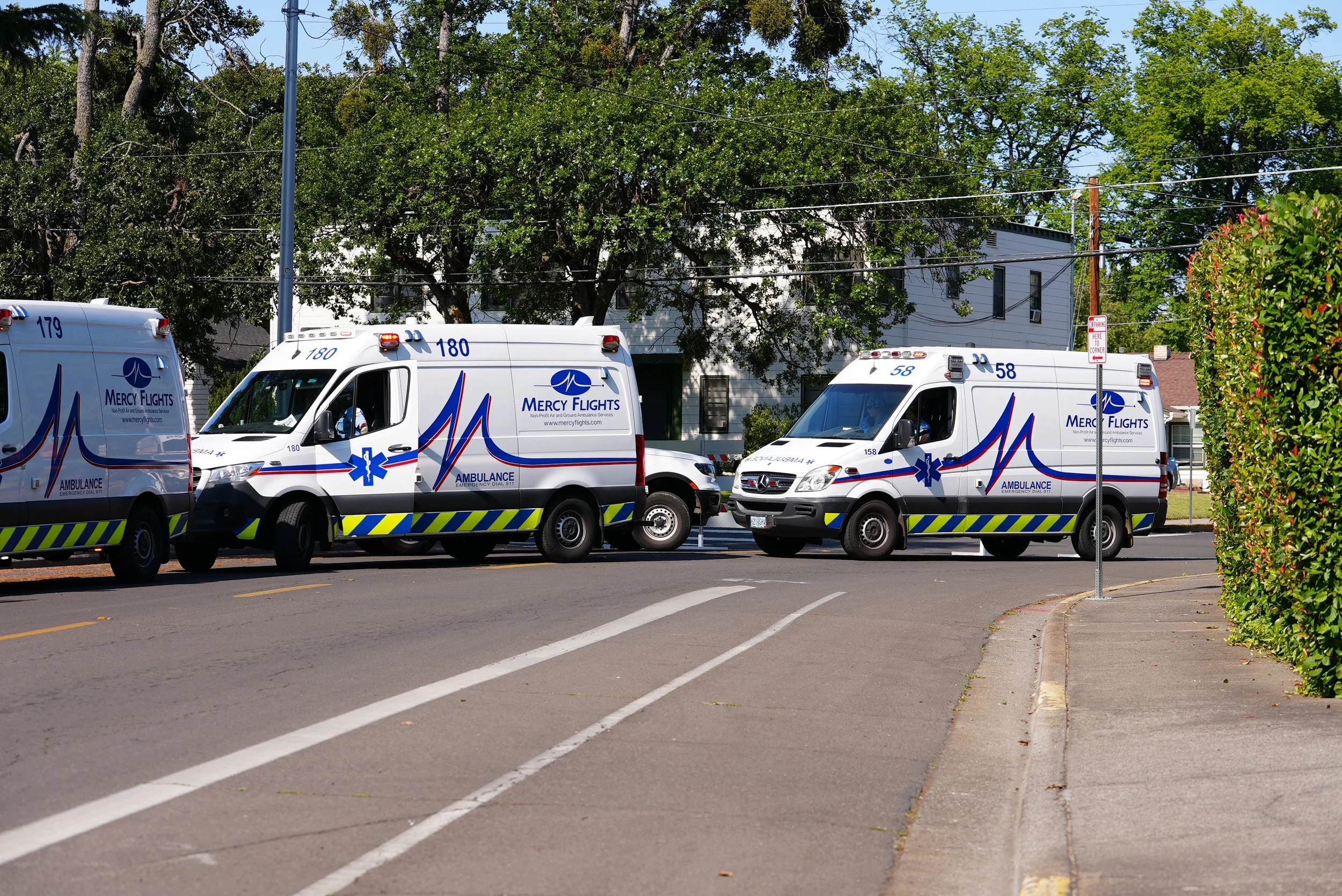 Three ambulance vehicles parked on a city street, with trees and a residential building in the background, and a mobility parking sign on the sidewalk.