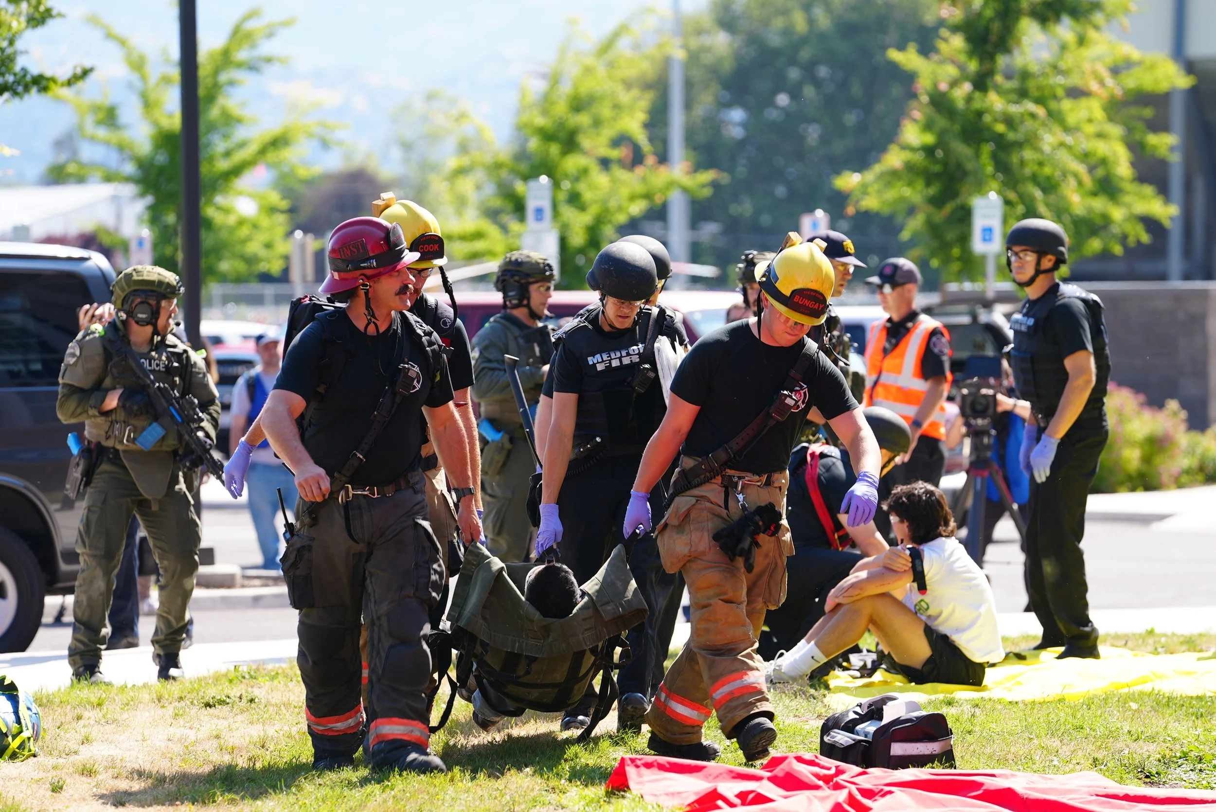 Emergency responders, including firefighters and police, attending to an injured person on a grassy area during a daytime incident.