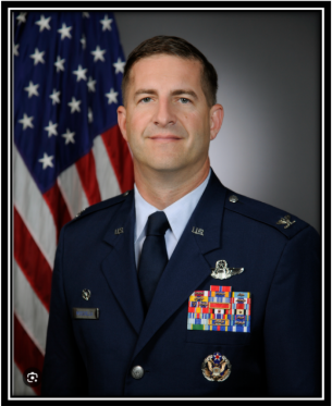 Portrait of a male military officer in uniform with medals, standing in front of an American flag.