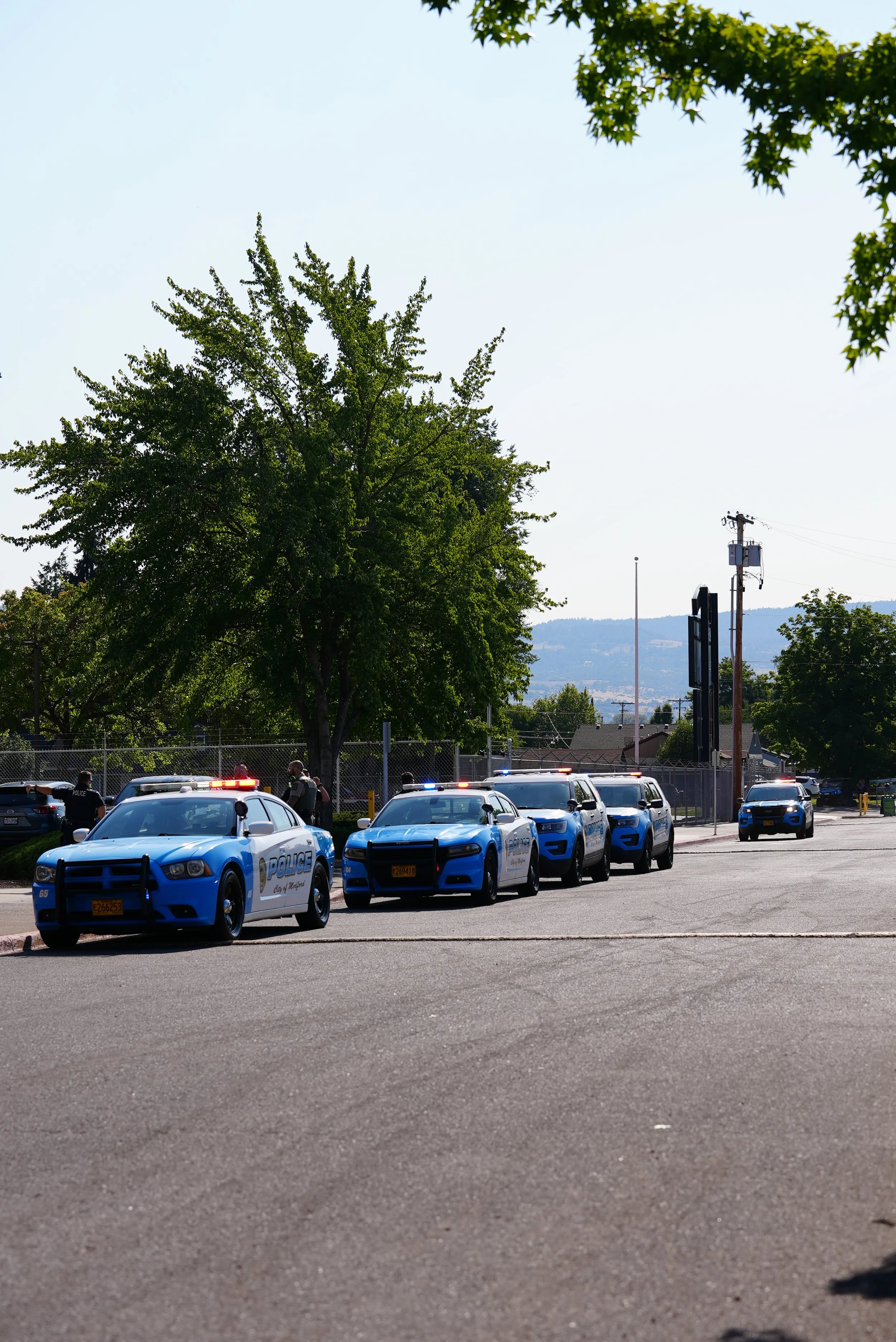 A parking lot with police cars and officers, surrounded by trees and a chain-link fence.