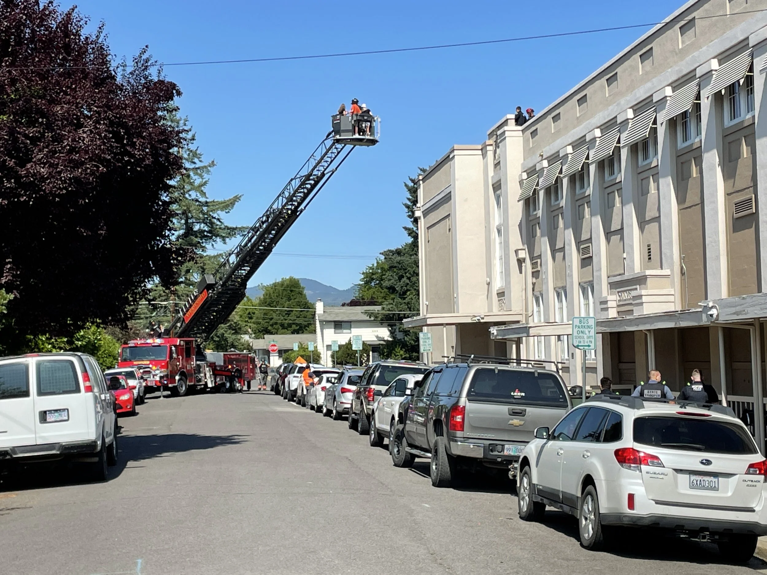 A fire truck with an extended ladder is parked on the street, with firefighters working at the top of the ladder. Several parked cars line the street, and a group of people, some in uniform, are gathered near the building on the right. The sky is cle