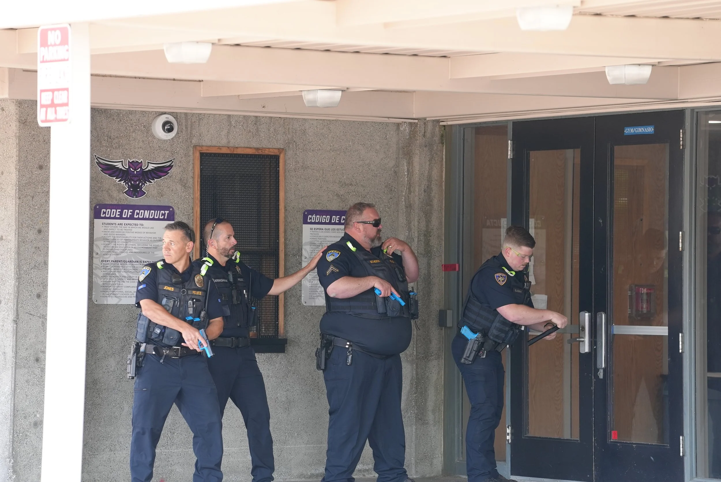Four police officers standing outside a building with glass doors, one is entering while the other three are standing nearby, one appears to be communicating with the others.